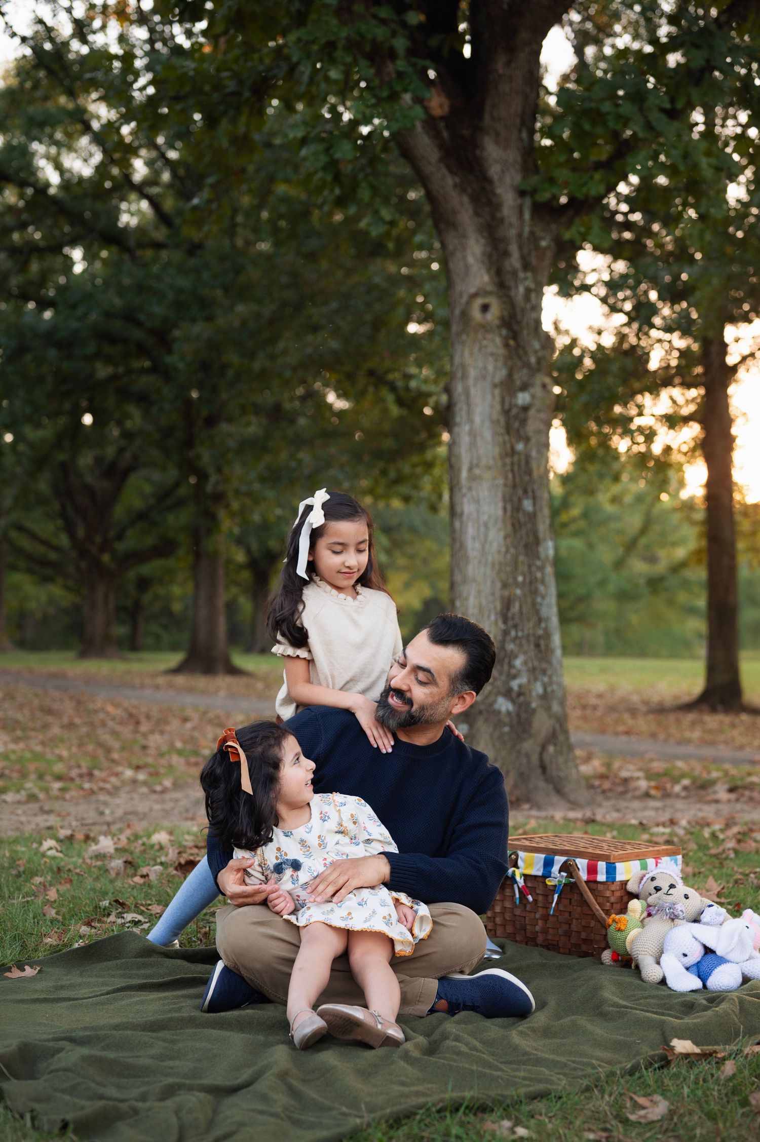 Dad sits on a blanket holding his younger daughter while the older sister leans on his shoulder, trees and picnic basket behind them.