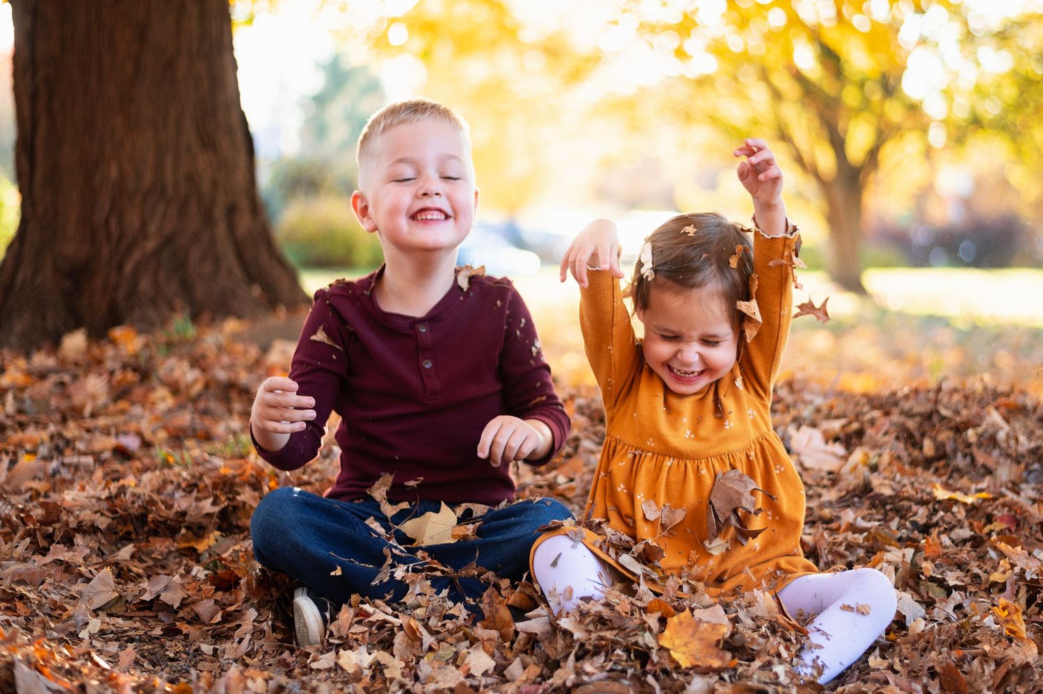 Two young children playing in a pile of autumn leaves, smiling and tossing them in the air under a golden tree.