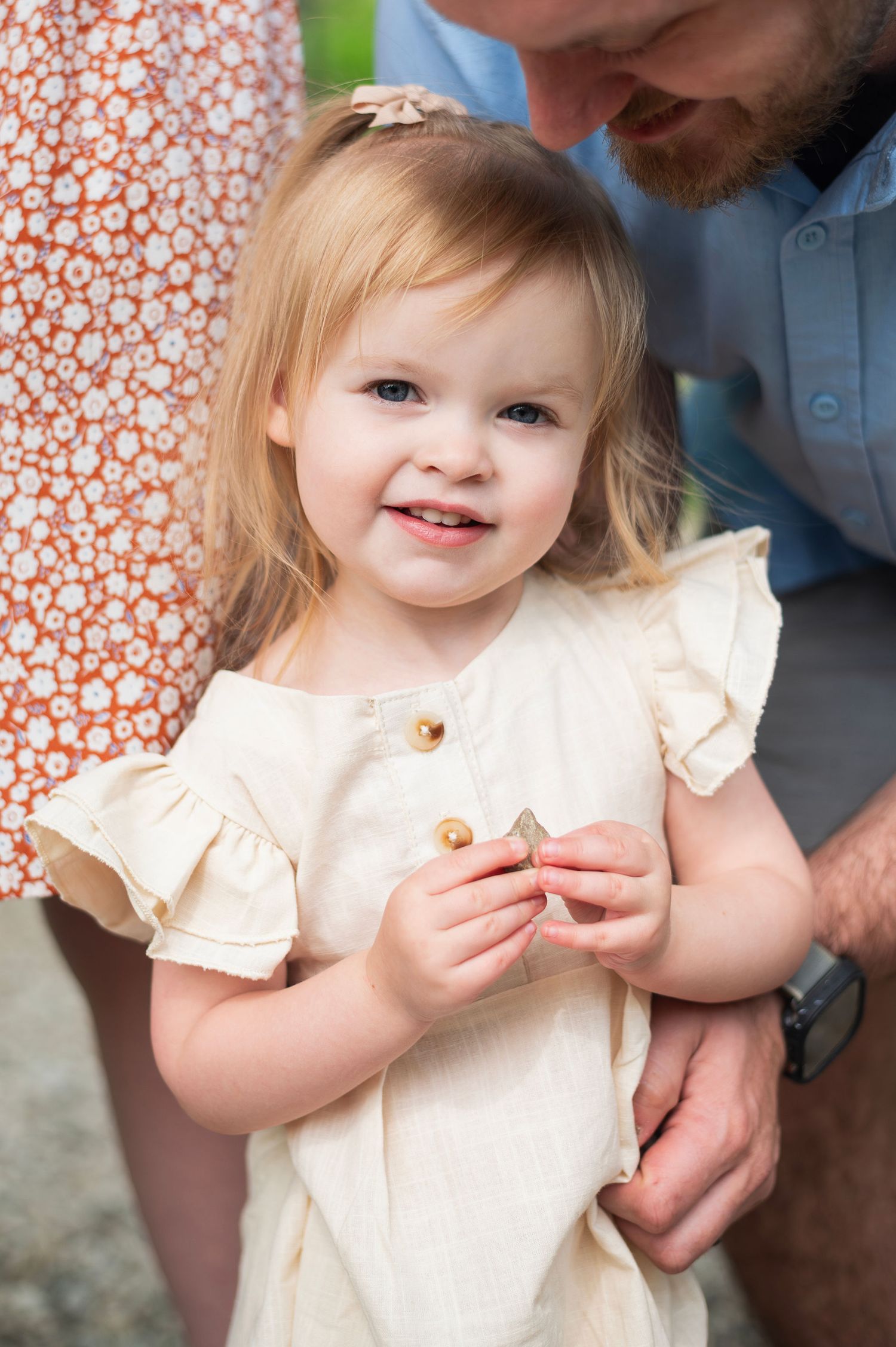 Toddler girl smiles up sweetly while holding a tiny treasure, surrounded by the gentle embrace of her parents.
