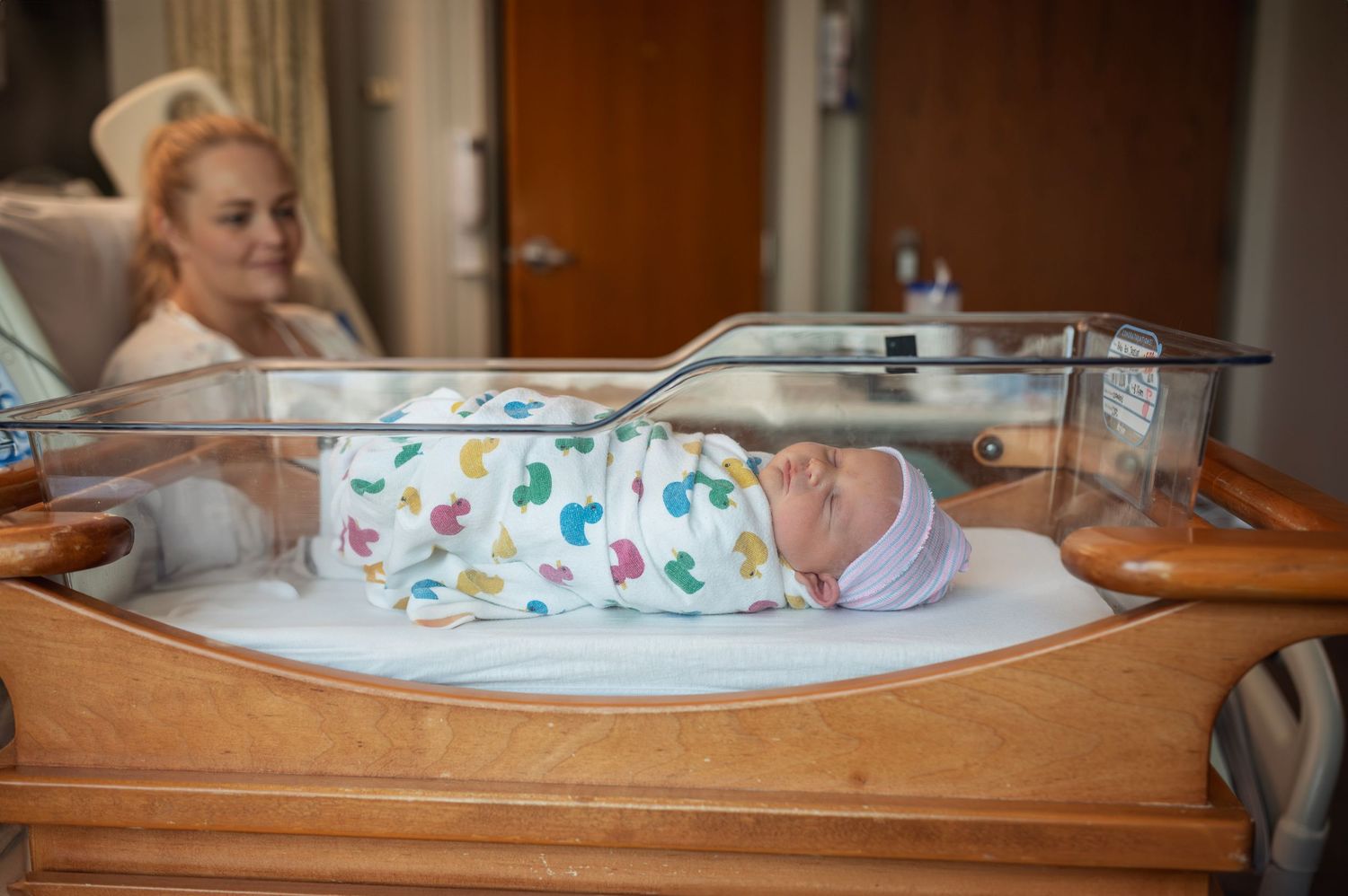 Mom watches lovingly as her newborn sleeps in bassinet wrapped in colorful duck swaddle during Fresh 48 session.