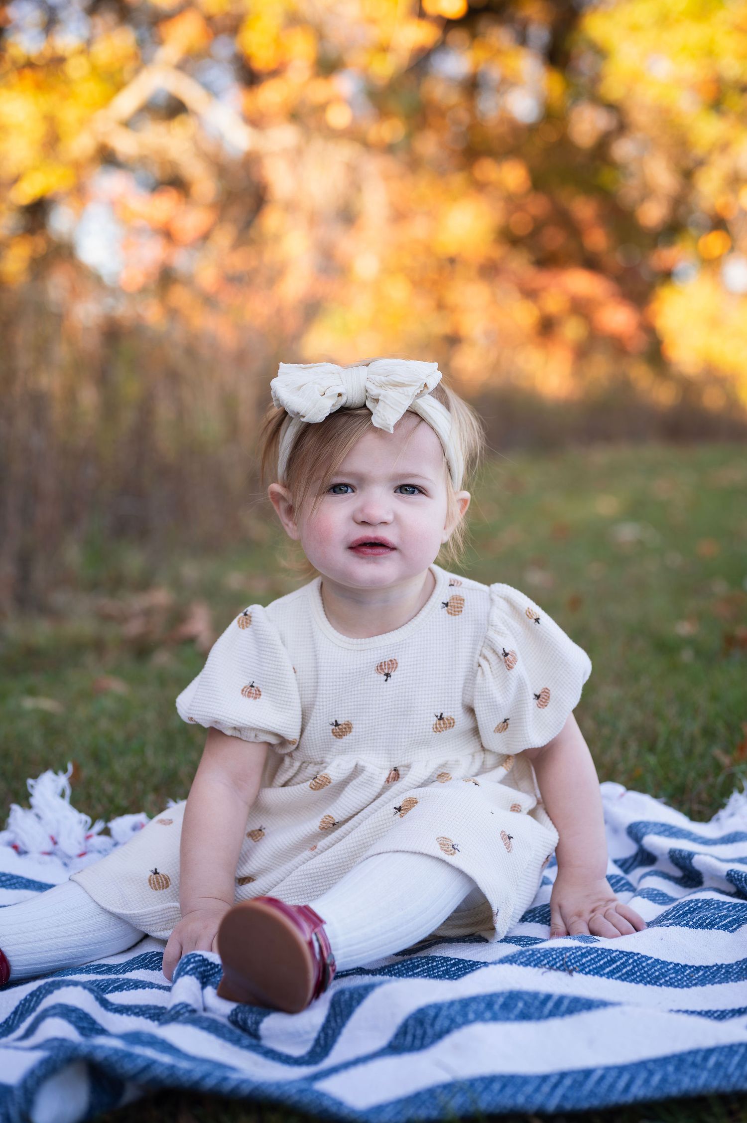 Toddler girl sits on a blanket in fall leaves, captured during a New Albany milestone session filled with warmth and whimsy.
