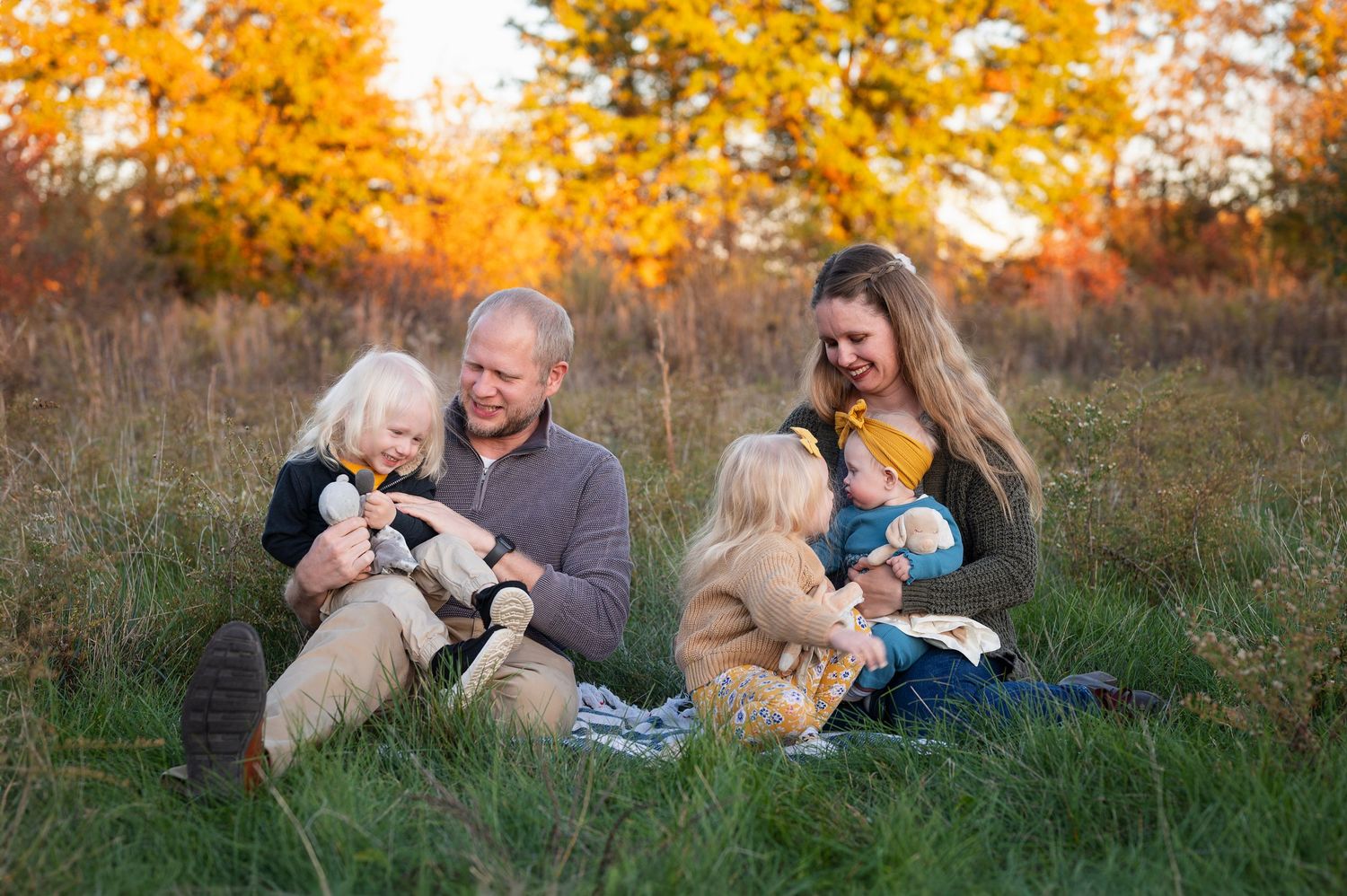Family of five sitting in a grassy field during golden hour, laughing together in cozy fall outfits.