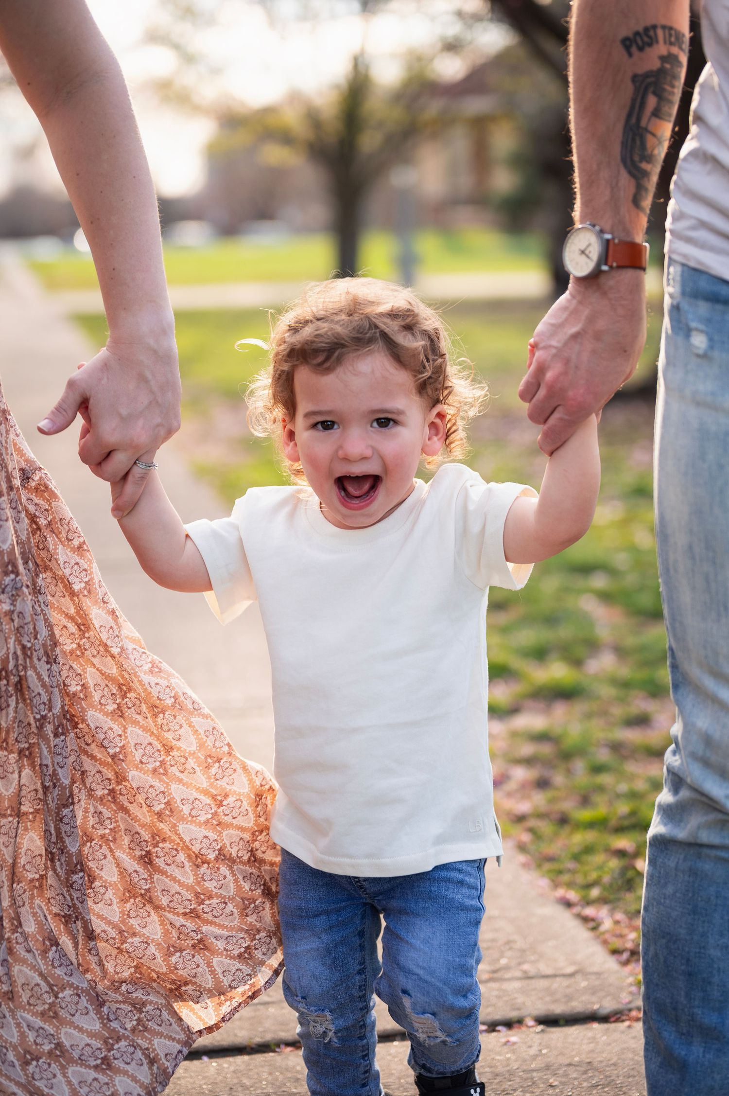 Toddler joyfully swinging between his parents’ hands on a sidewalk at Goodale Park in Columbus, Ohio.