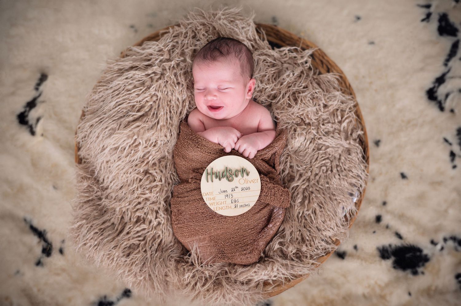 Smiling newborn boy swaddled in brown wrap, lying in a fur-lined basket holding a wooden name sign.