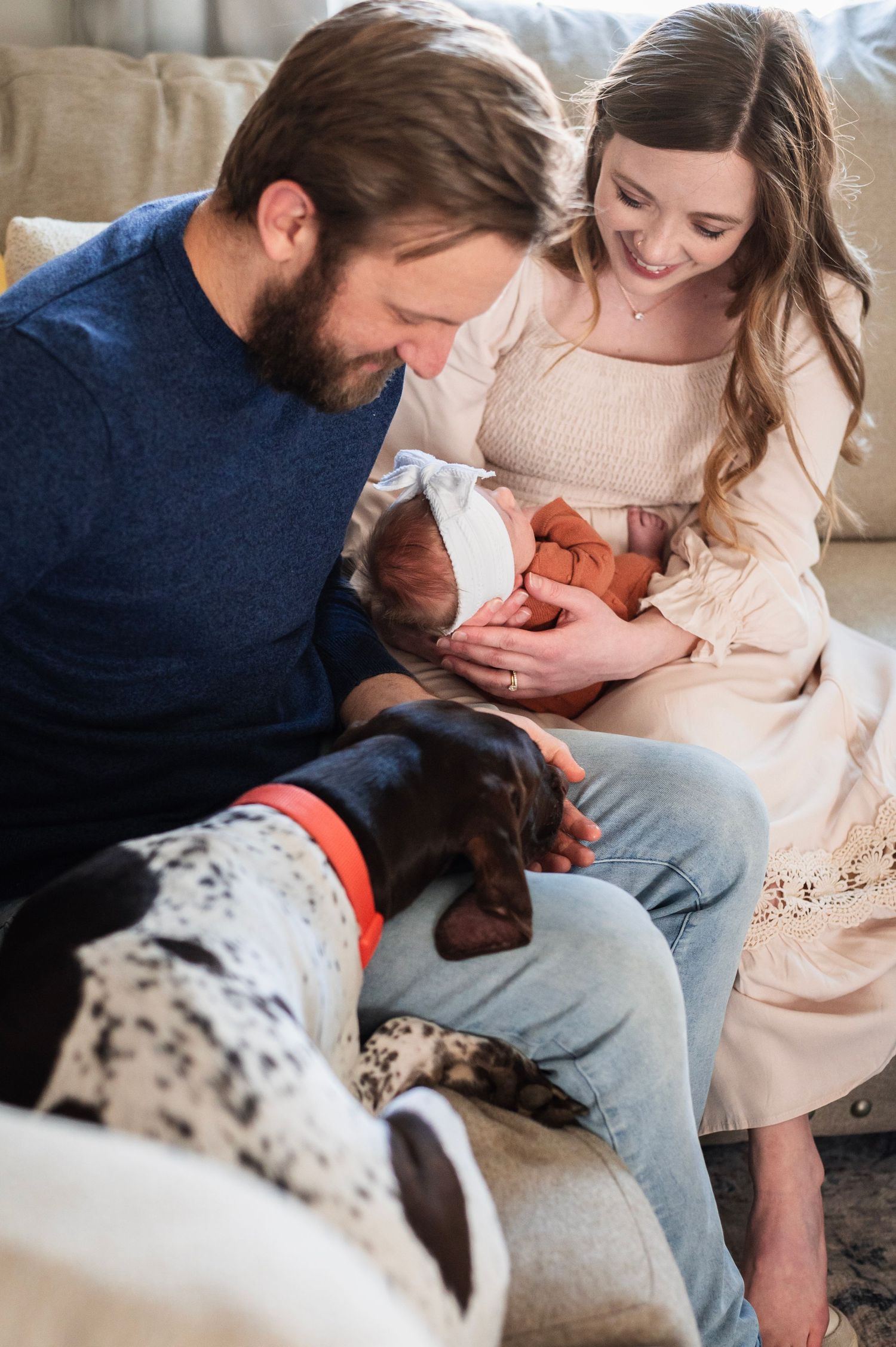 Family cuddles their newborn on the couch while their spotted dog sniffs gently, capturing love in a cozy in-home moment.