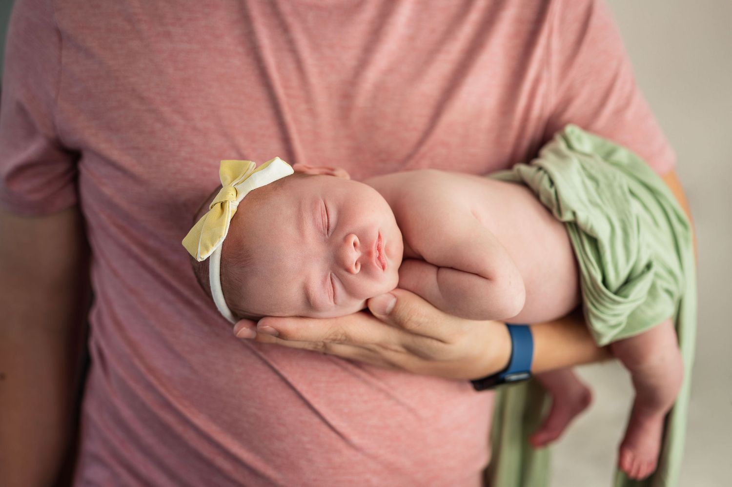 Newborn girl in a soft wrap and yellow bow, cradled in dad’s hands. Westerville newborn photographer.