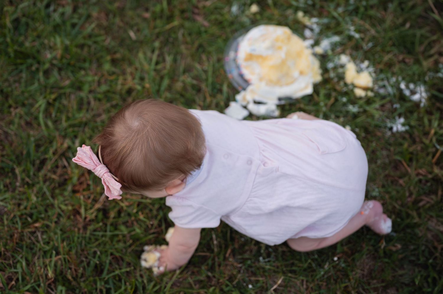 Overhead view of baby crawling in grass after a cake smash, frosting-covered cake scattered nearby.