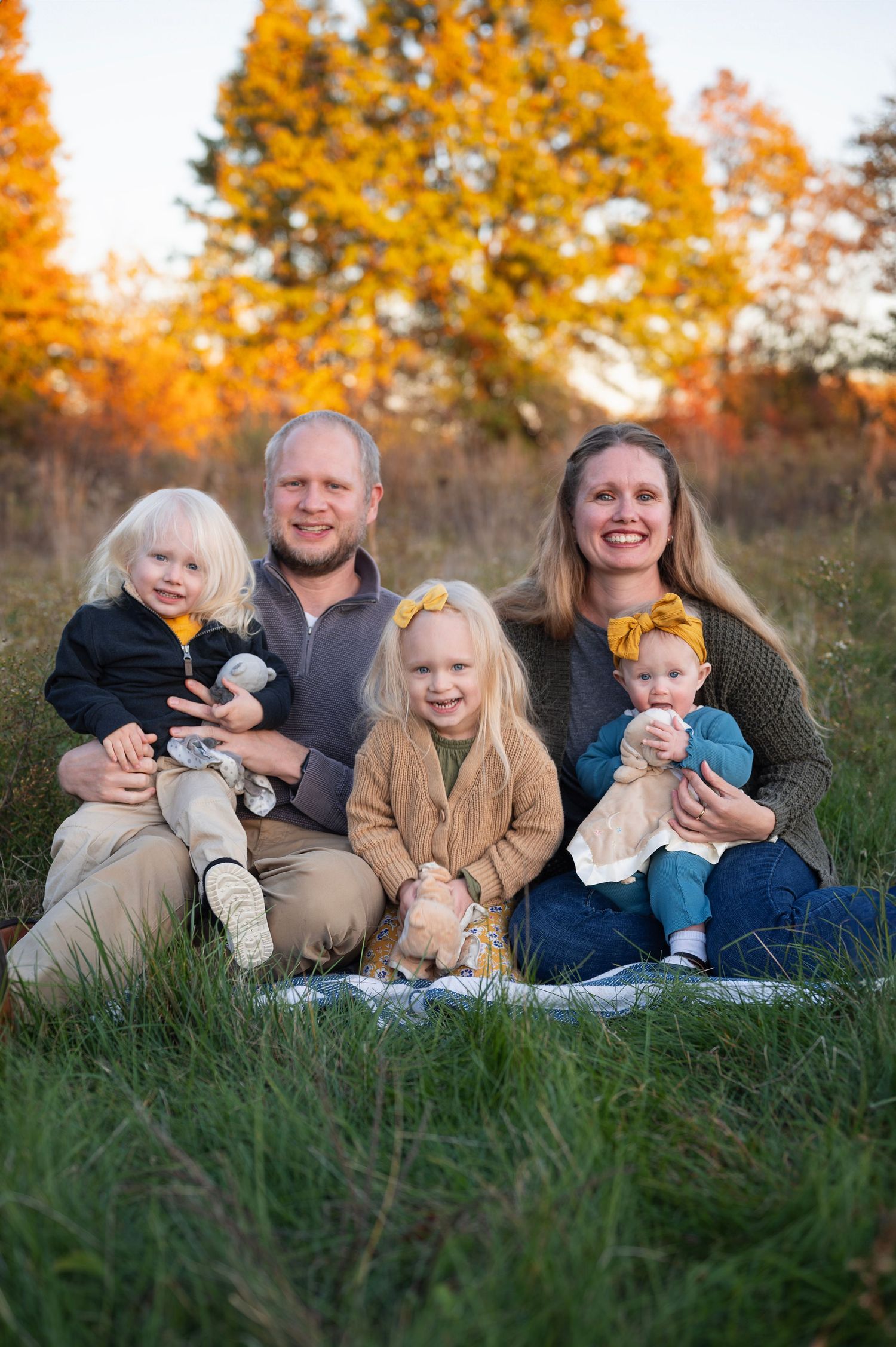 Smiling family of five sitting in a grassy field with golden autumn trees in the background, wearing cozy fall outfits with yellow accents