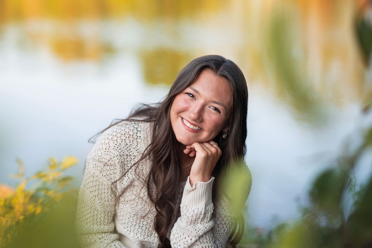 Teen girl in cream sweater and white pants sitting on a rock by the water, smiling in soft fall light. Sharon Woods