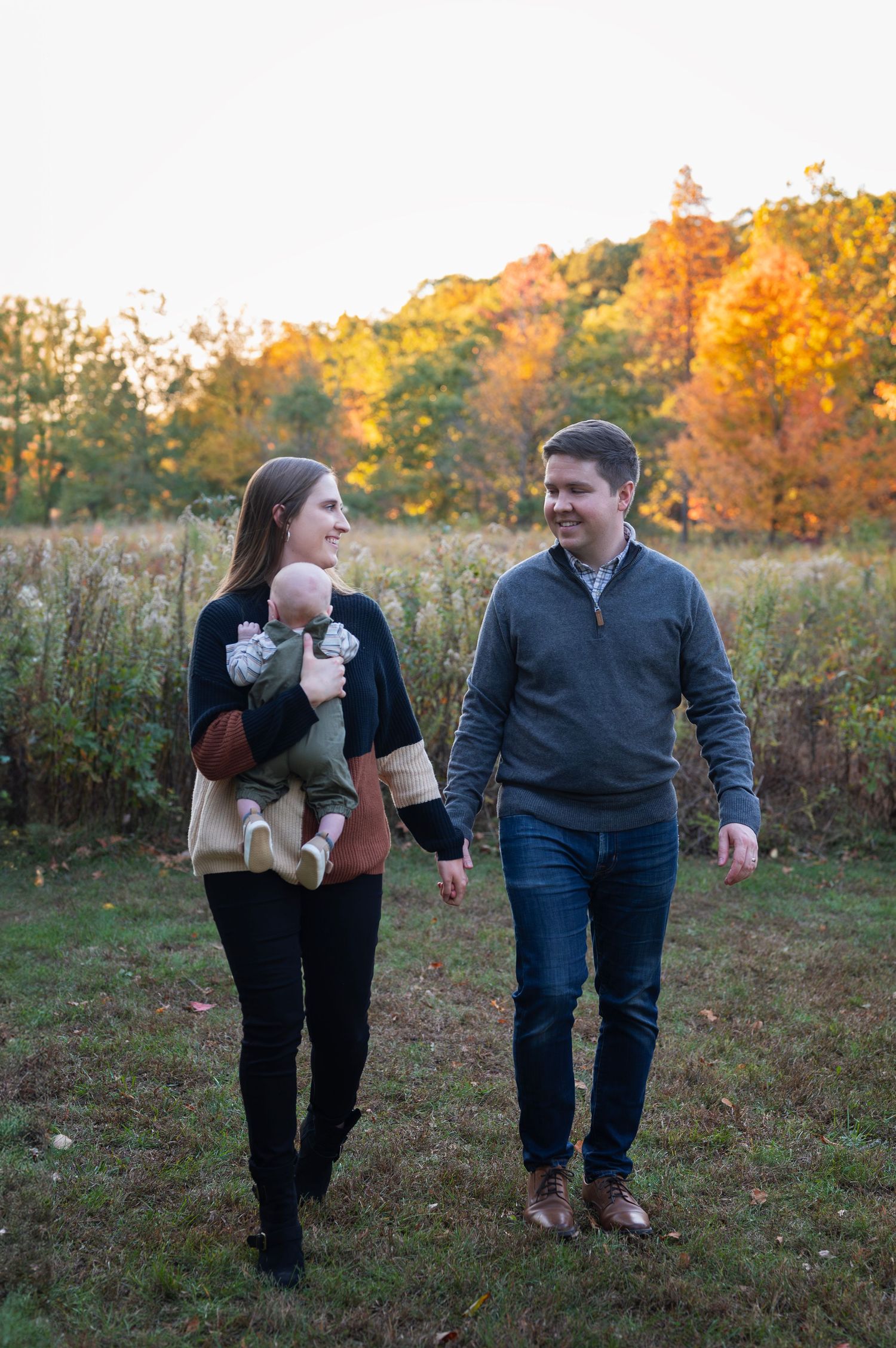 Parents holding hands while mom carries baby, walking through a meadow with golden autumn trees in the background. Highbanks Metro Park