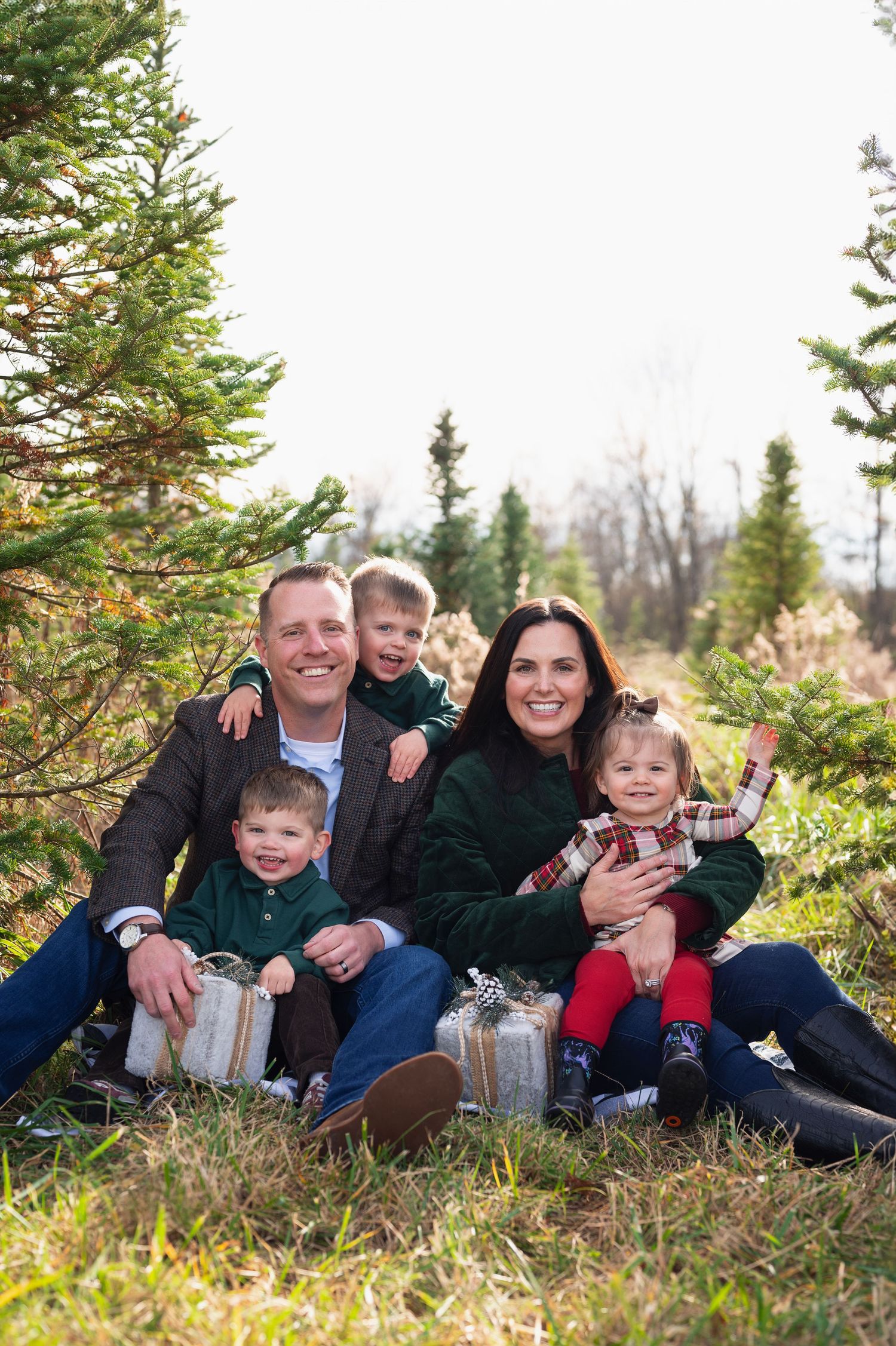 Family of five sitting among Christmas trees, holding wrapped presents and smiling for the camera.