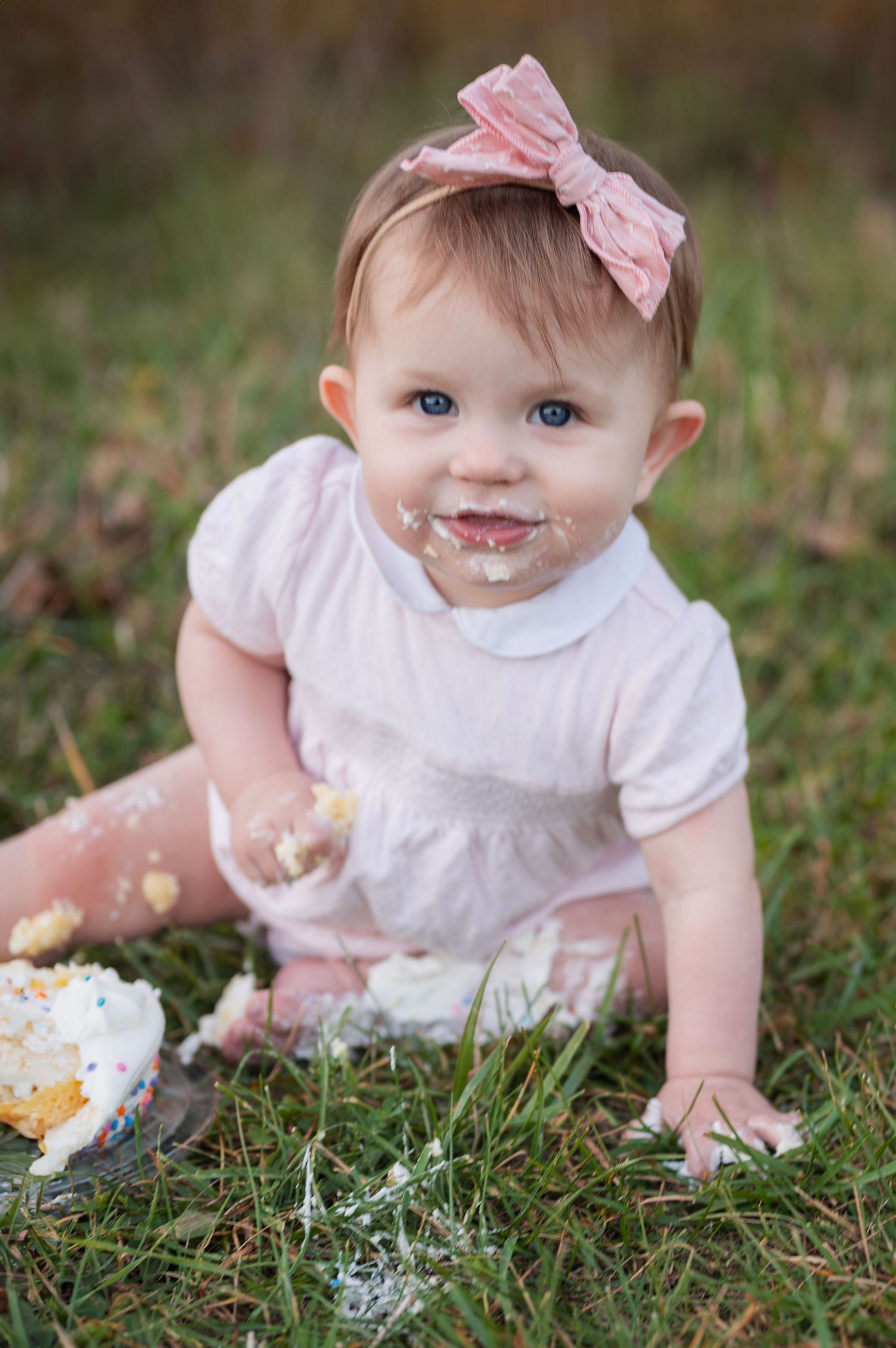 One-year-old smiles sweetly with frosting on her face after a joyful outdoor cake smash session.