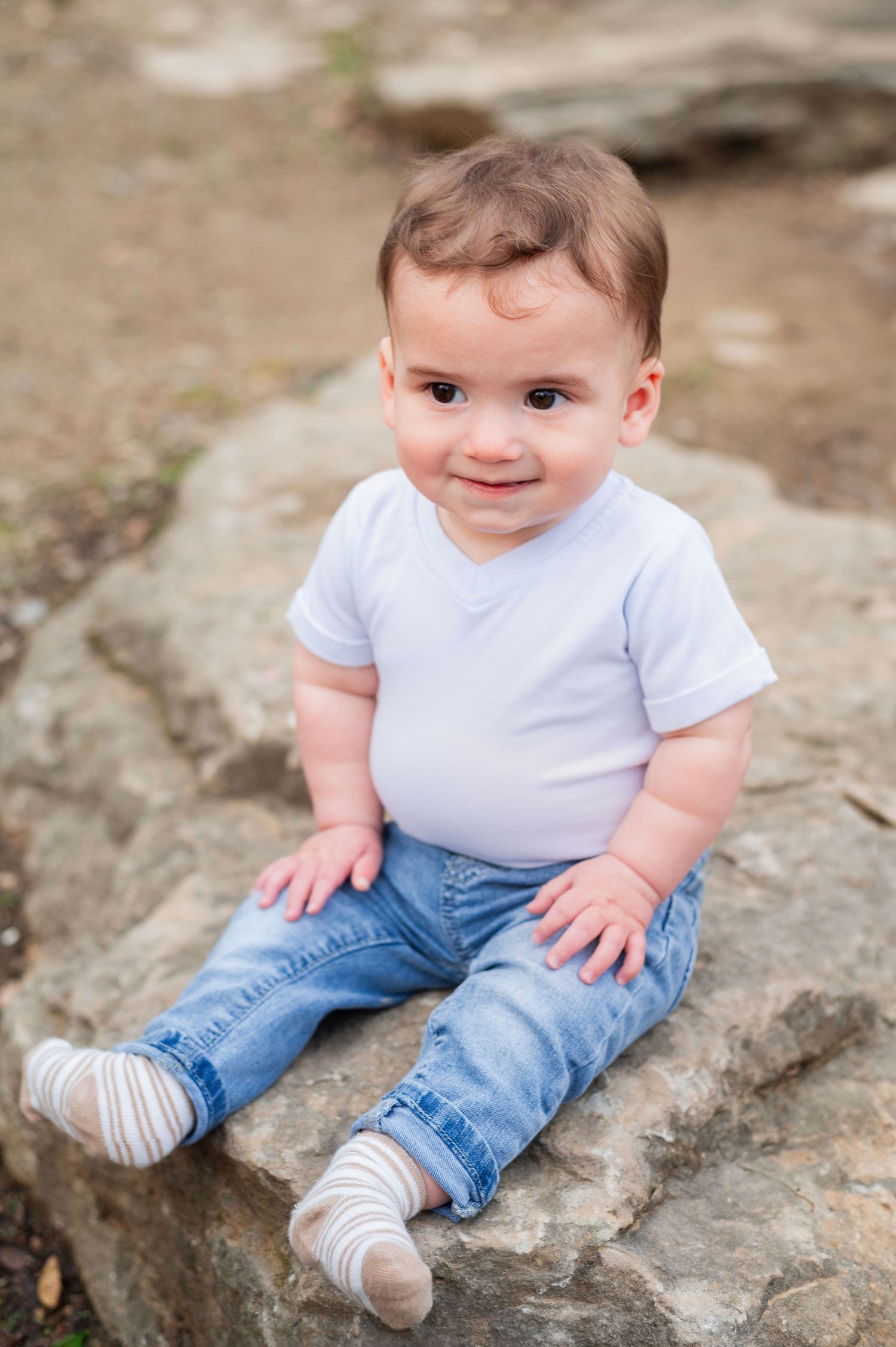 Sweet baby boy sits on a rock, smiling shyly during a milestone photography session filled with curiosity and charm.
