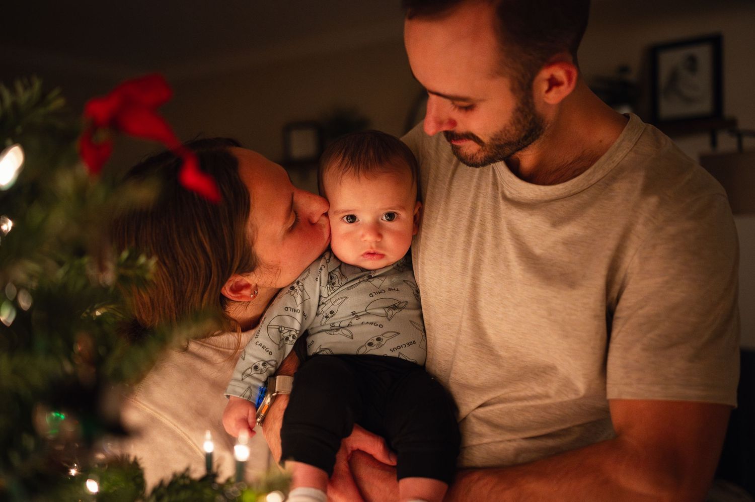 Mom kisses baby by the Christmas tree while dad holds them both, glowing with soft holiday light.