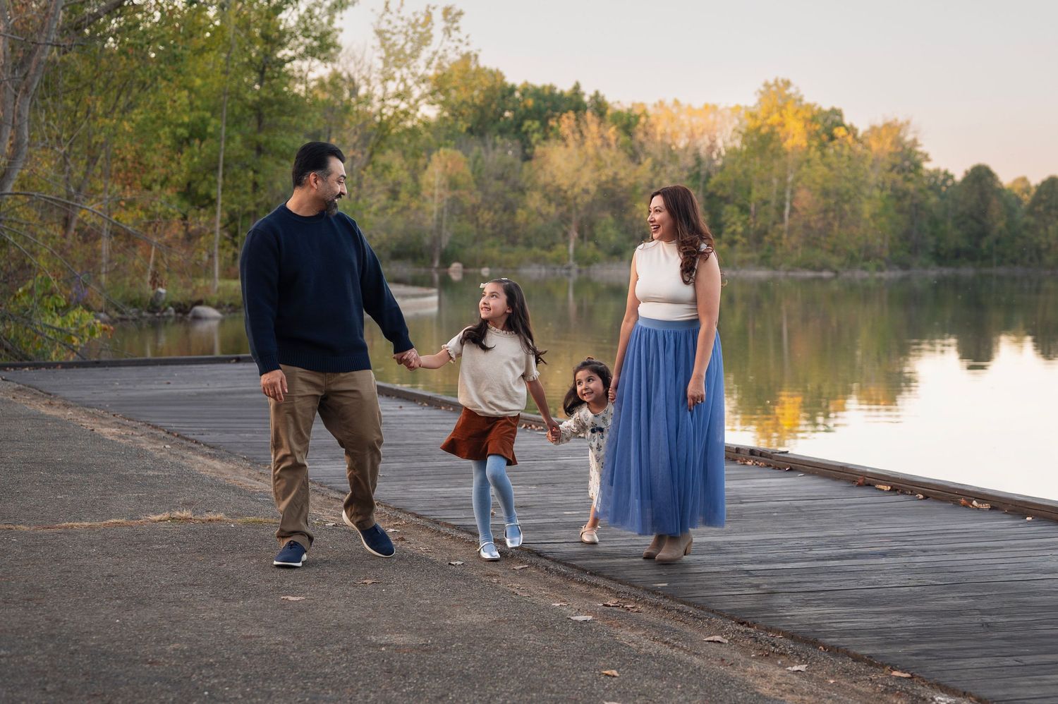Westerville family photographer captures a family of four holding hands and walking along a lakeside dock at sunset in early fall.