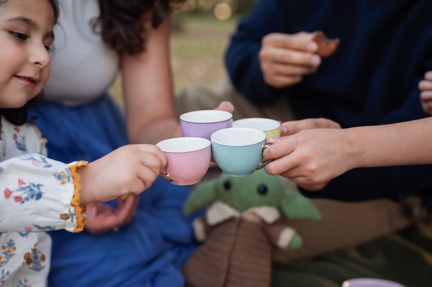 Close-up of pastel toy tea cups clinked together at a family tea party, with a crocheted Grogu doll in the background.