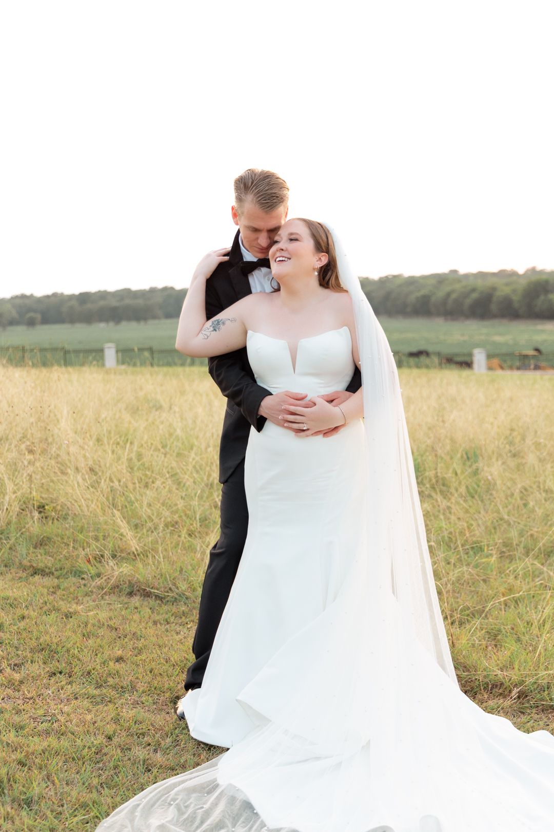 Groom embraces smiling bride in strapless gown with long veil in an open golden field at dusk.
