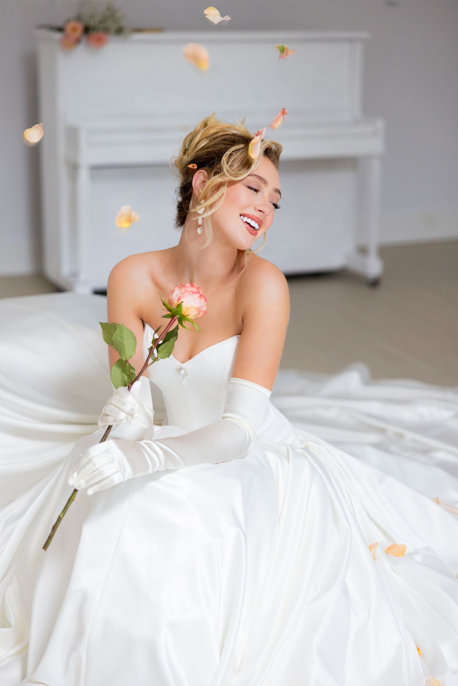 Joyful bride in white strapless gown and gloves holds a pink rose as petals fall around her near a white piano.