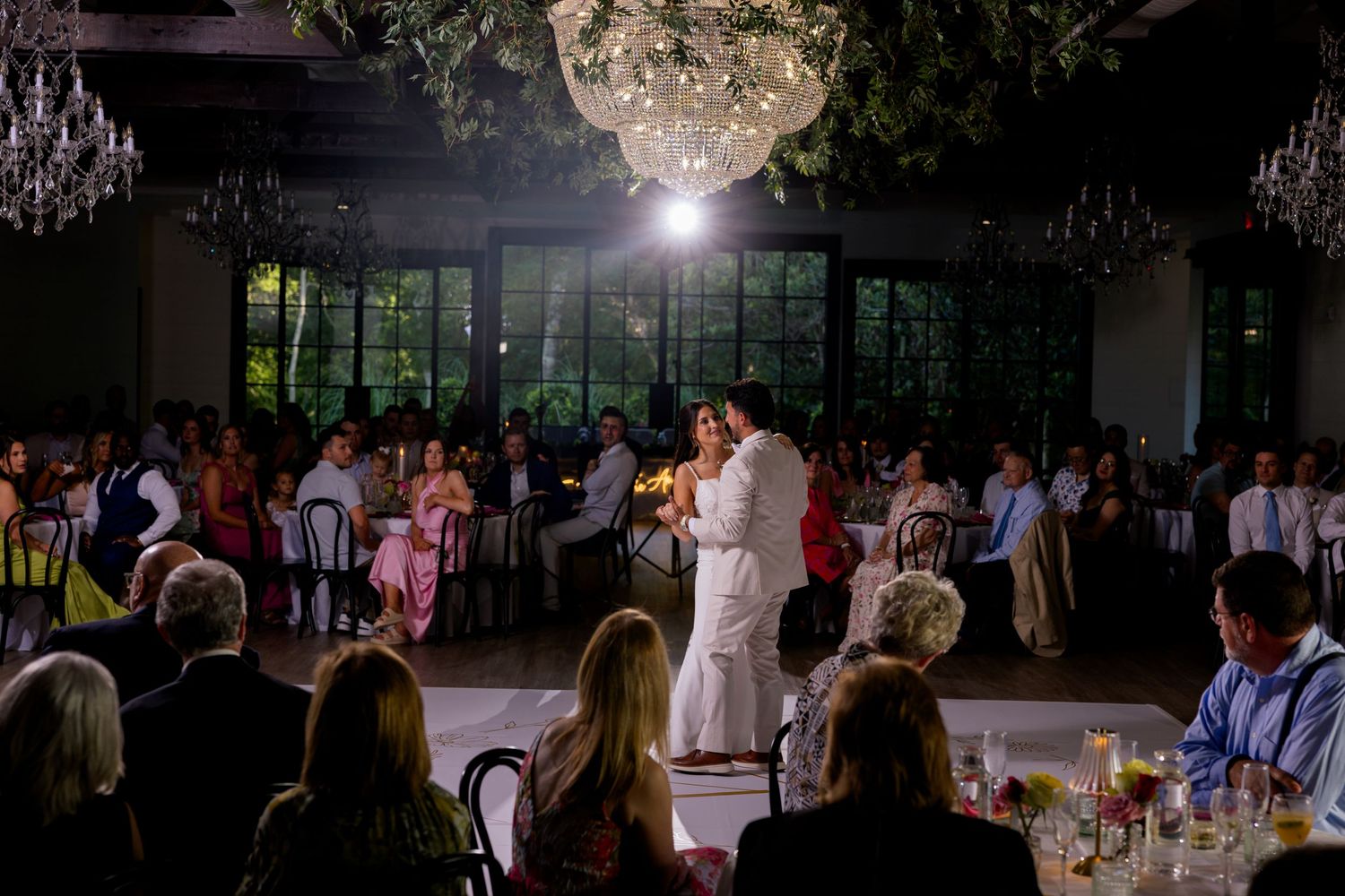 Couple shares first dance at elegant wedding reception with chandeliers and guests seated at decorated tables.