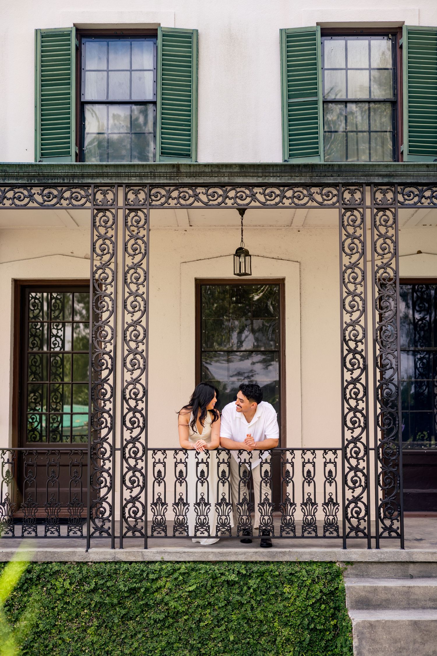 Couple sharing a romantic moment on an ornate wrought iron porch of a historic cream building with green shutters.