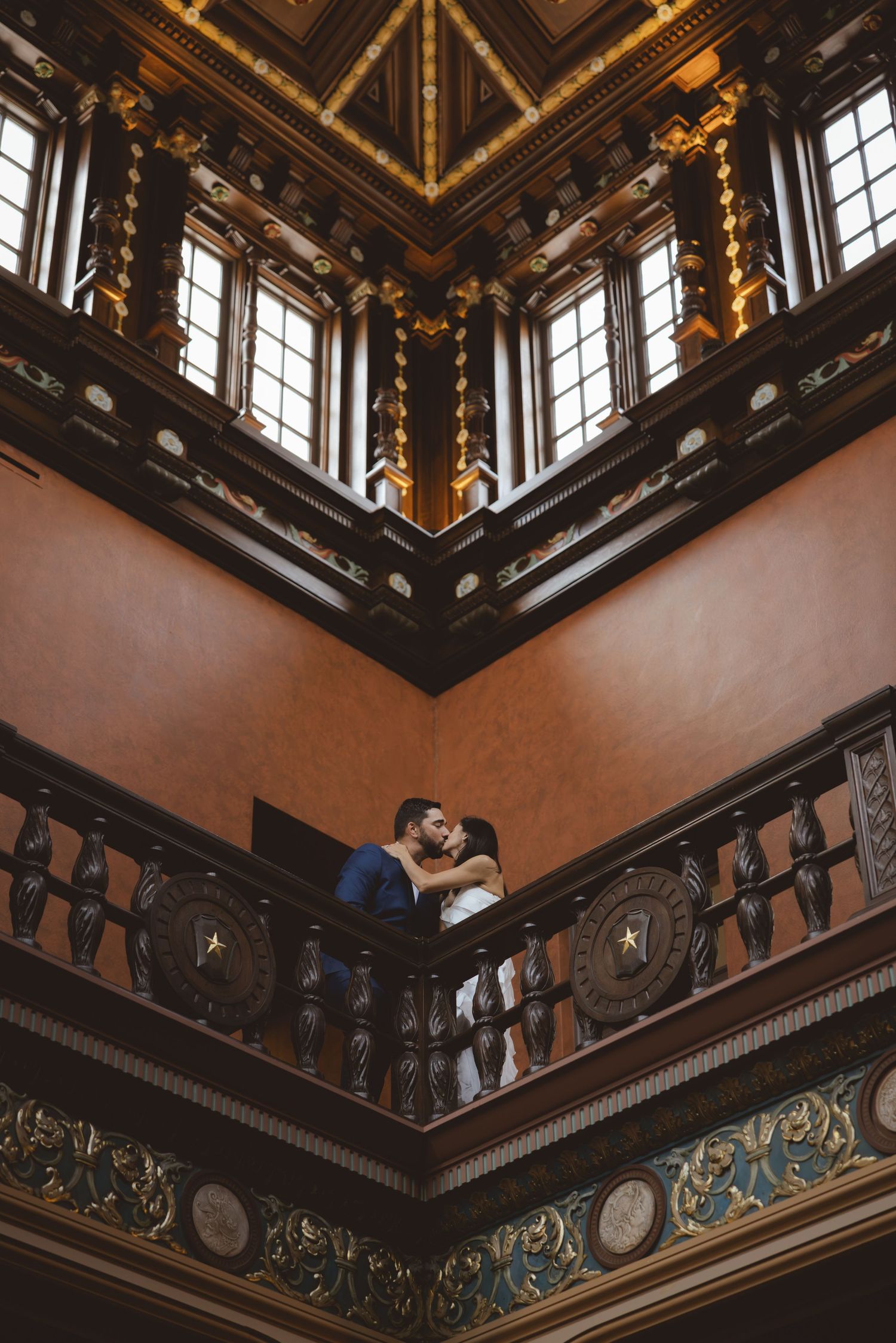Couple sharing a romantic kiss on an ornate balcony with carved railings inside a grand historic building.