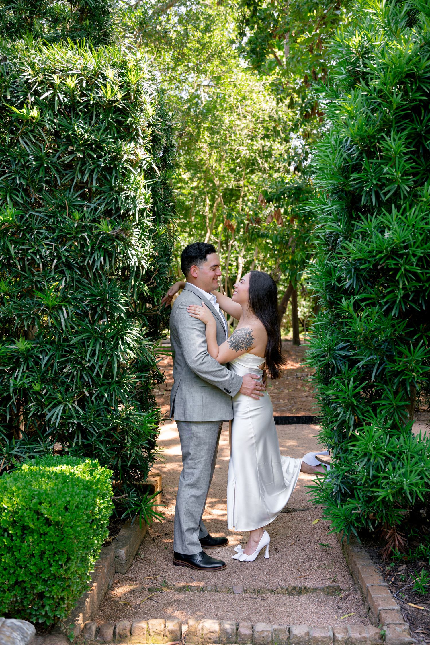 A couple in wedding attire embraces on a garden path surrounded by lush green hedges and trees.