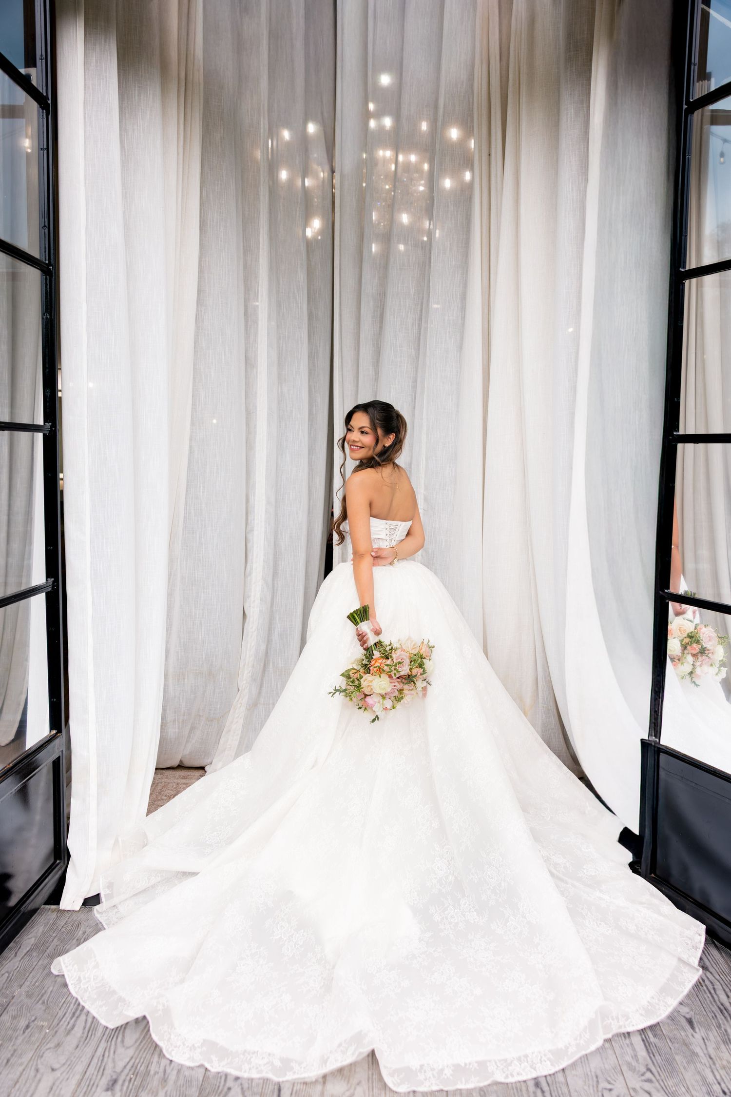 Bride in a strapless ball gown with long train holds bouquet, posing by sheer curtains under chandelier.