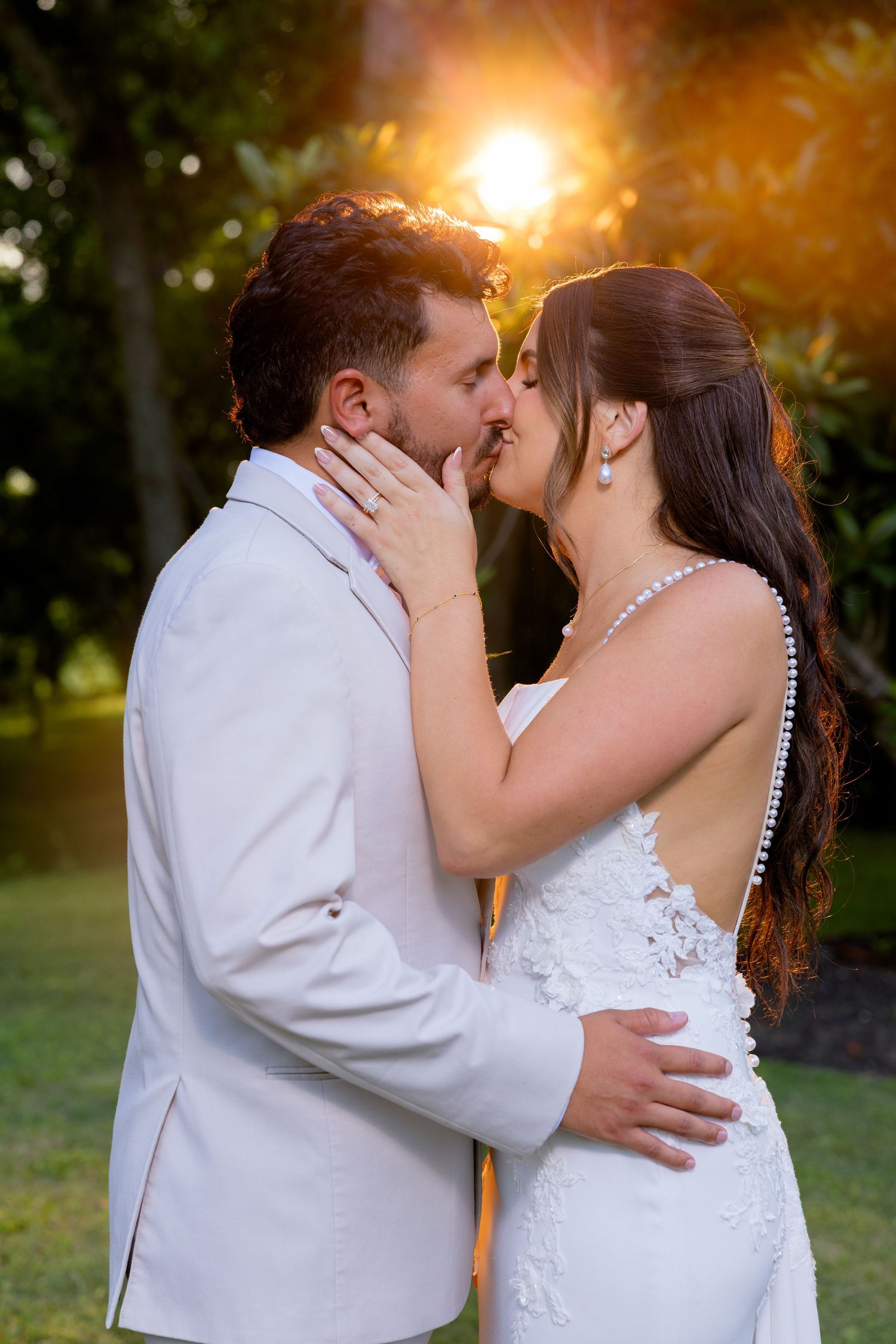 Bride and groom share a romantic sunset kiss outdoors, wearing white wedding attire with golden light glowing behind them.