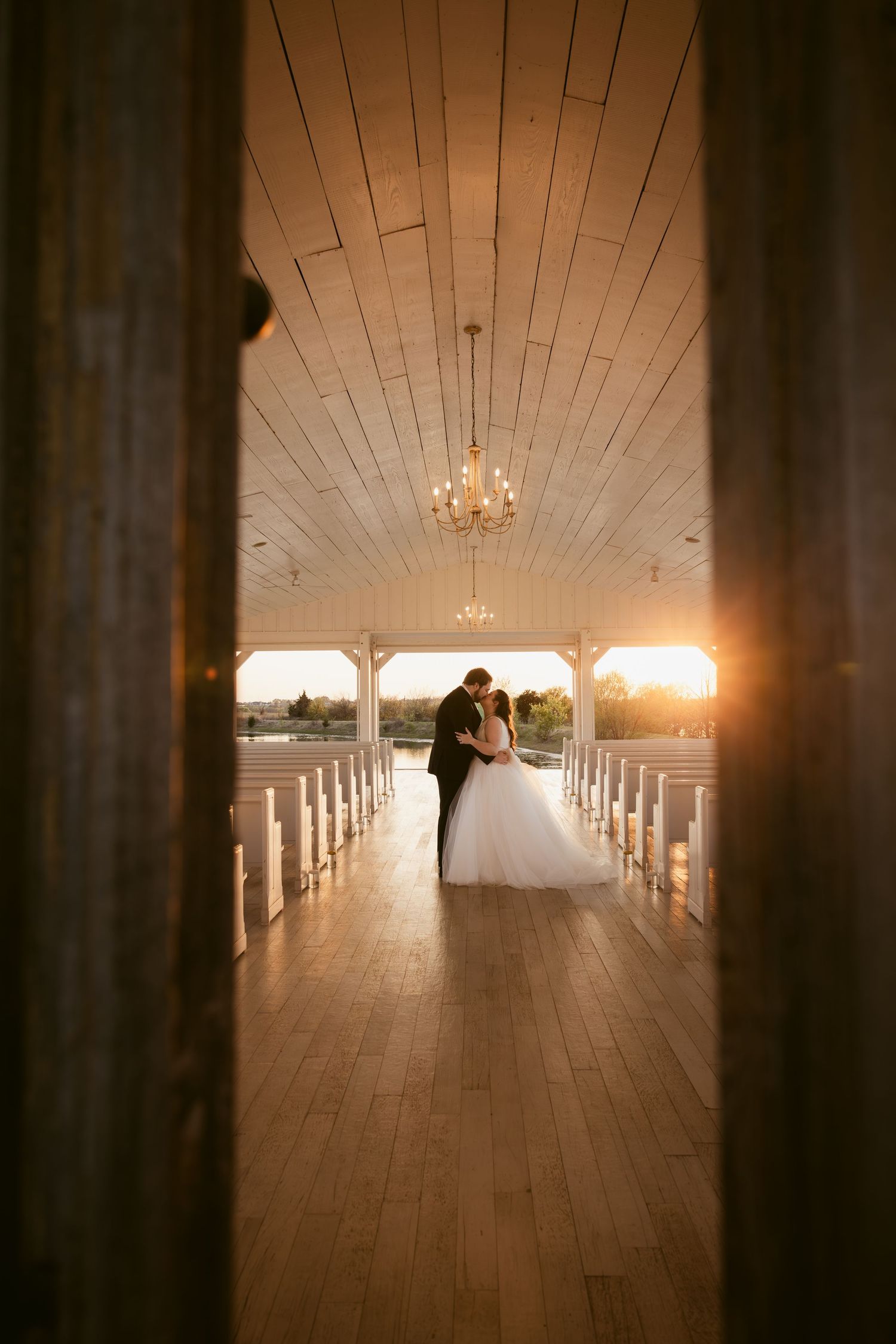 Bride and groom share a romantic kiss at sunset inside an elegant rustic chapel with wooden floors and chandelier.