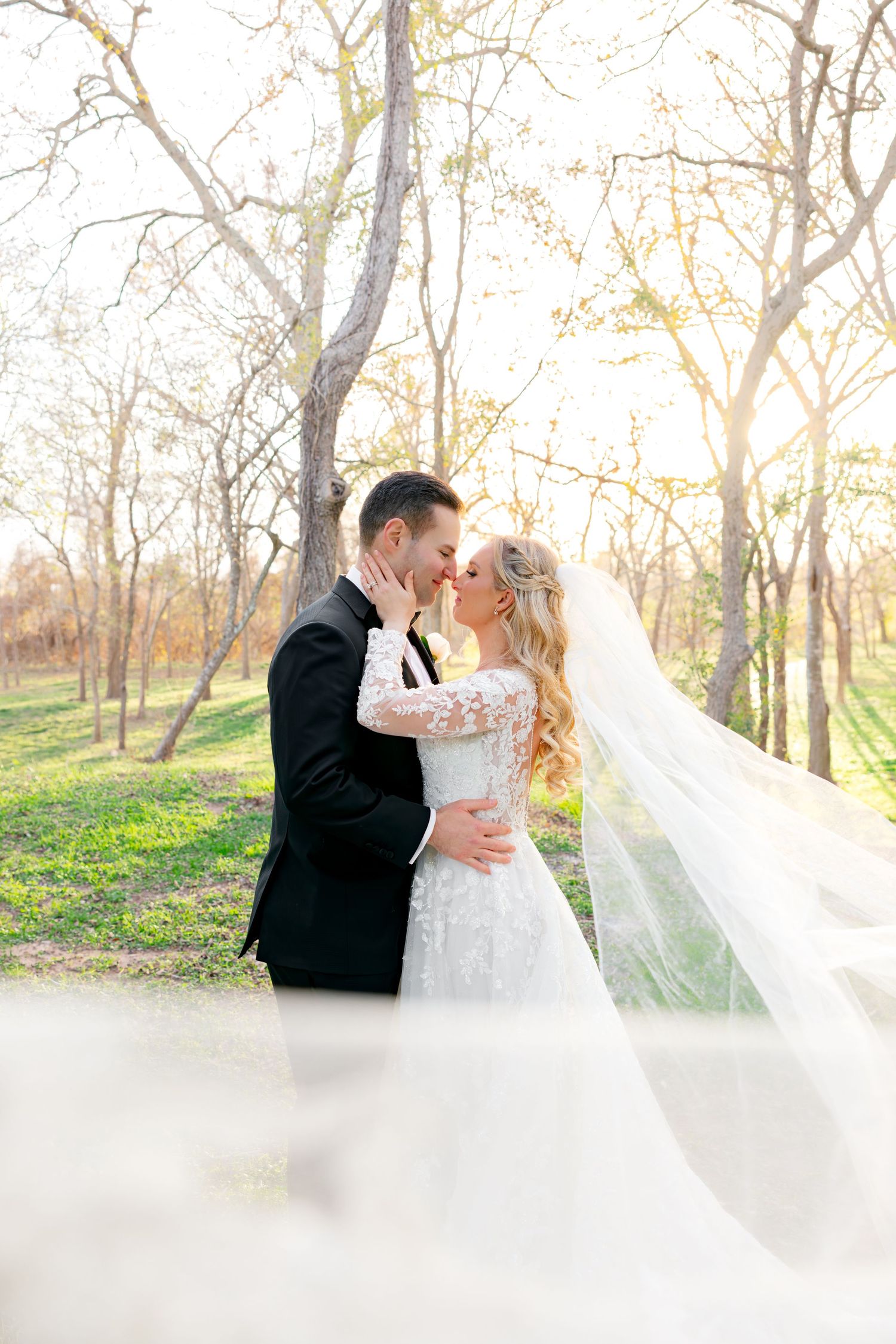 Bride and groom share a romantic moment in a sunlit forest, her veil flowing gracefully as they lean in for a kiss.