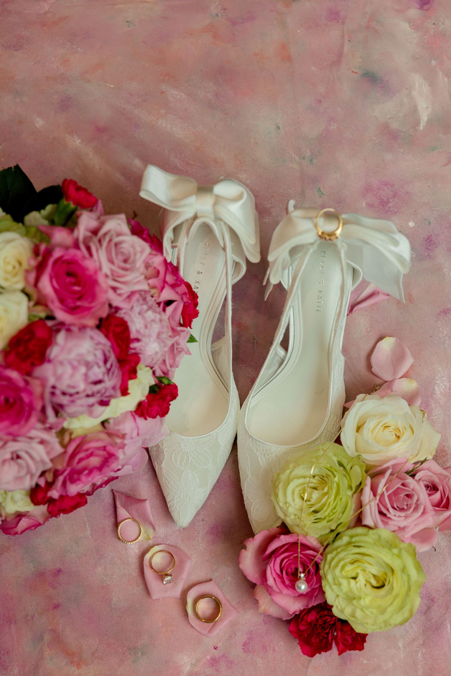 White satin bow heels surrounded by pink roses, gold rings, and a bridal bouquet on a pink background.