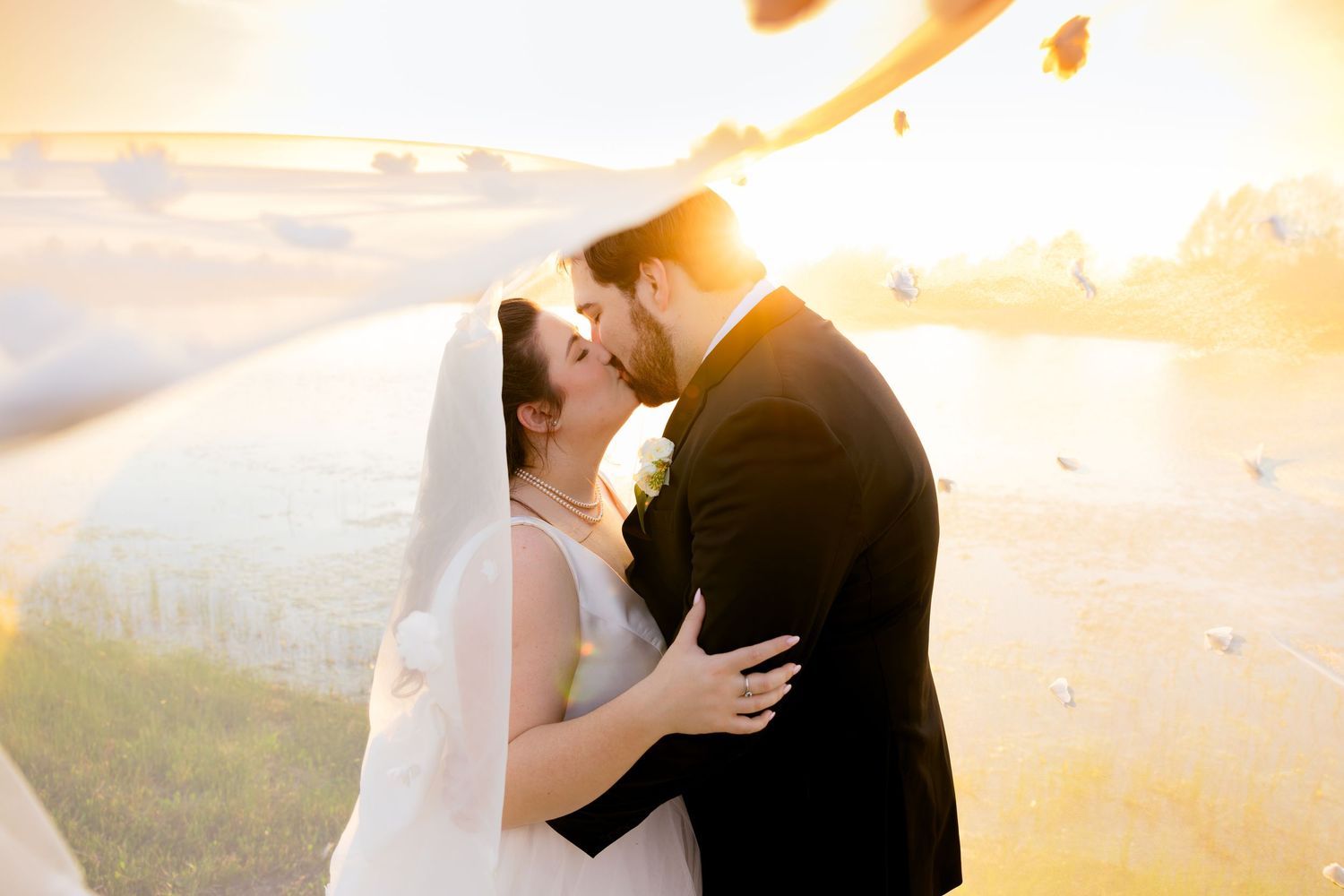 Bride and groom sharing a romantic kiss at golden hour sunset, with soft bokeh and flower petals floating in the air.