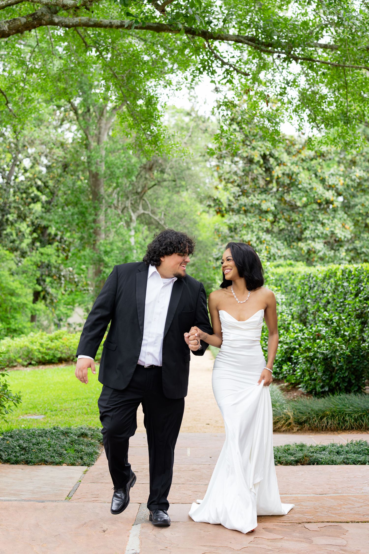 A bride and groom walk hand-in-hand through a lush green garden, smiling at each other in wedding attire.