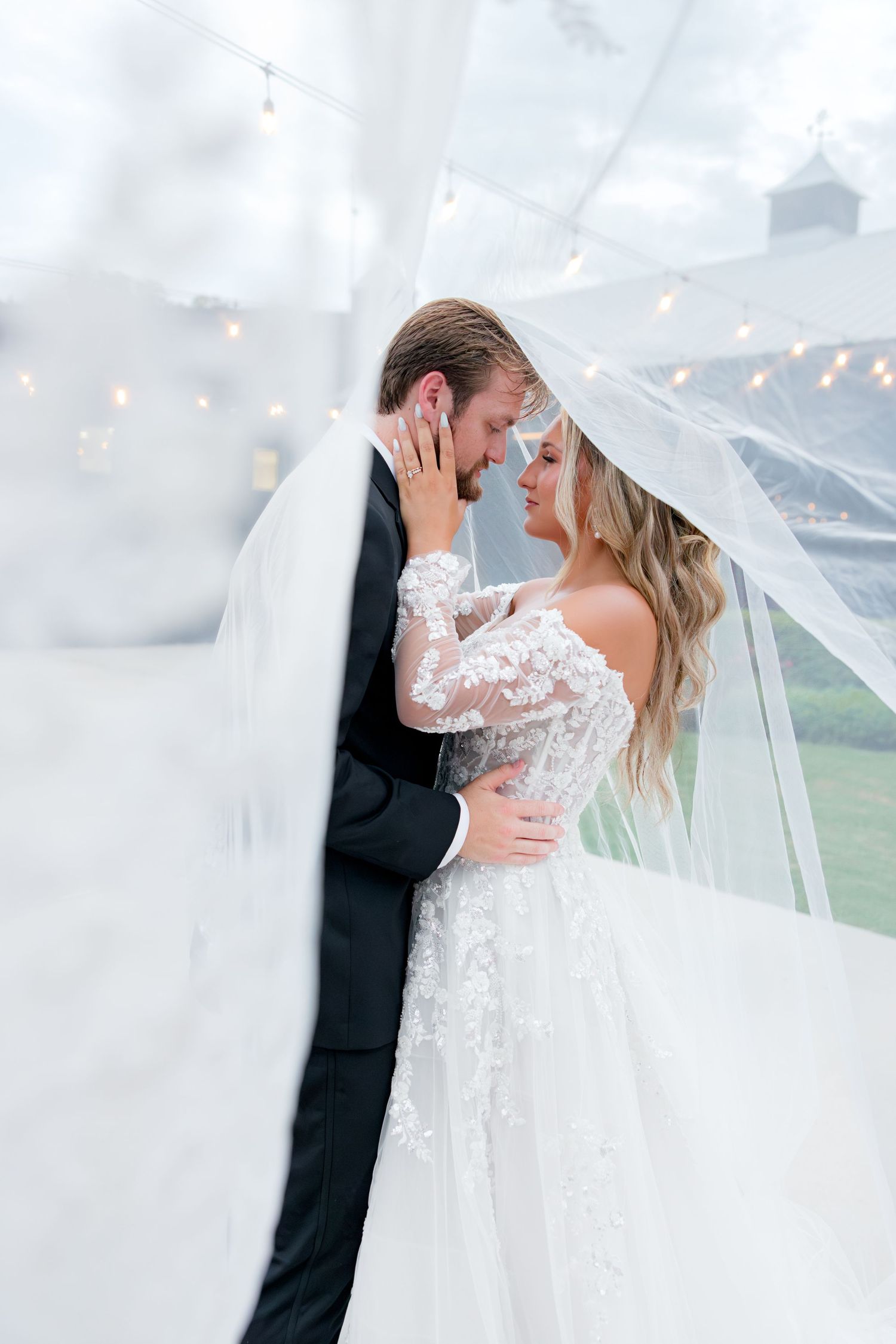 Bride and groom share romantic moment under flowing veil at elegant outdoor wedding venue with string lights.