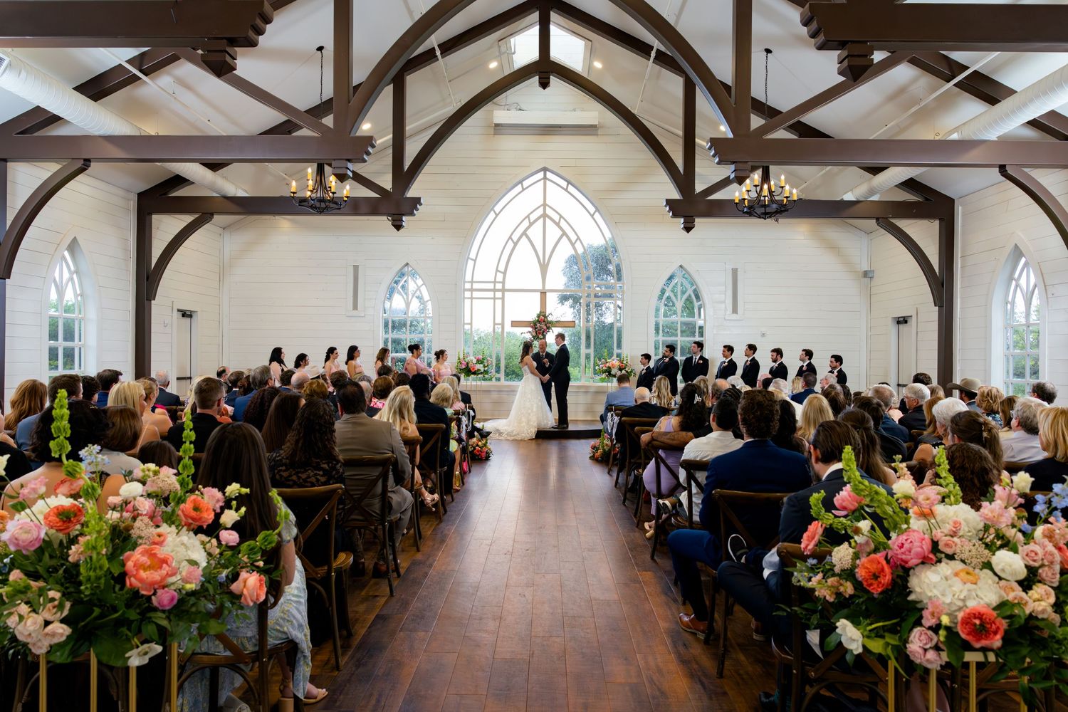 Elegant white chapel wedding ceremony with gothic windows, floral arrangements, and guests seated in wooden pews.