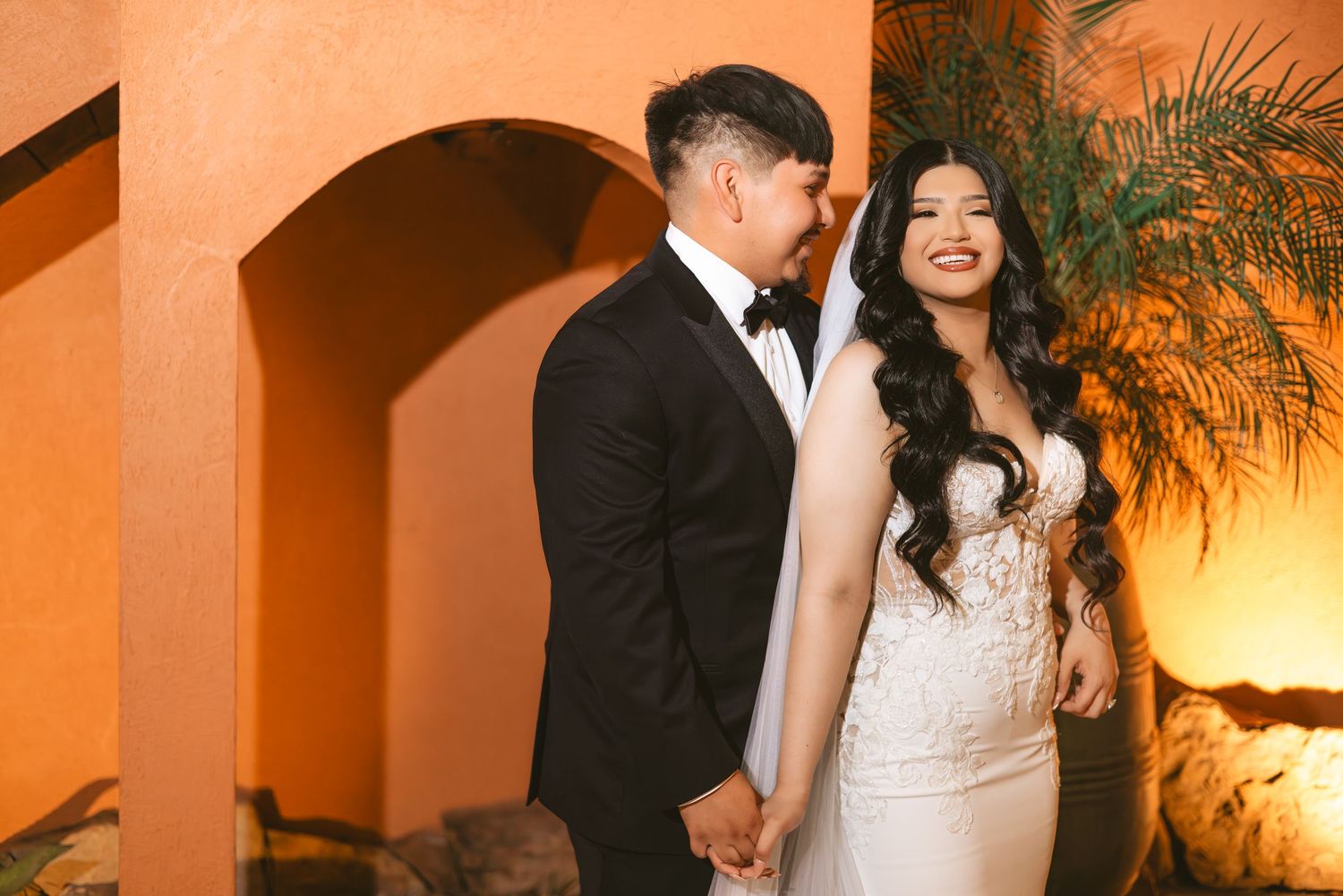 Elegant couple poses at night, bride in fitted lace gown and groom in tuxedo, holding hands under warm amber lighting.