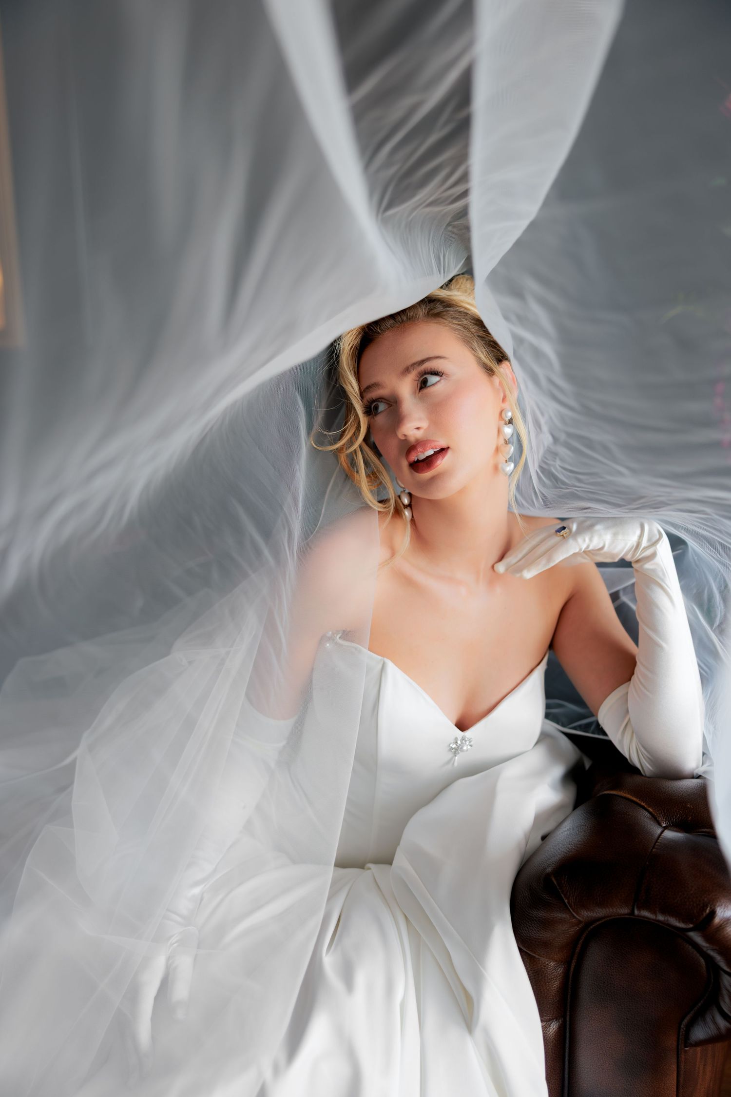 A bride in a white strapless gown and gloves sits gracefully as her flowing veil billows dramatically around her.