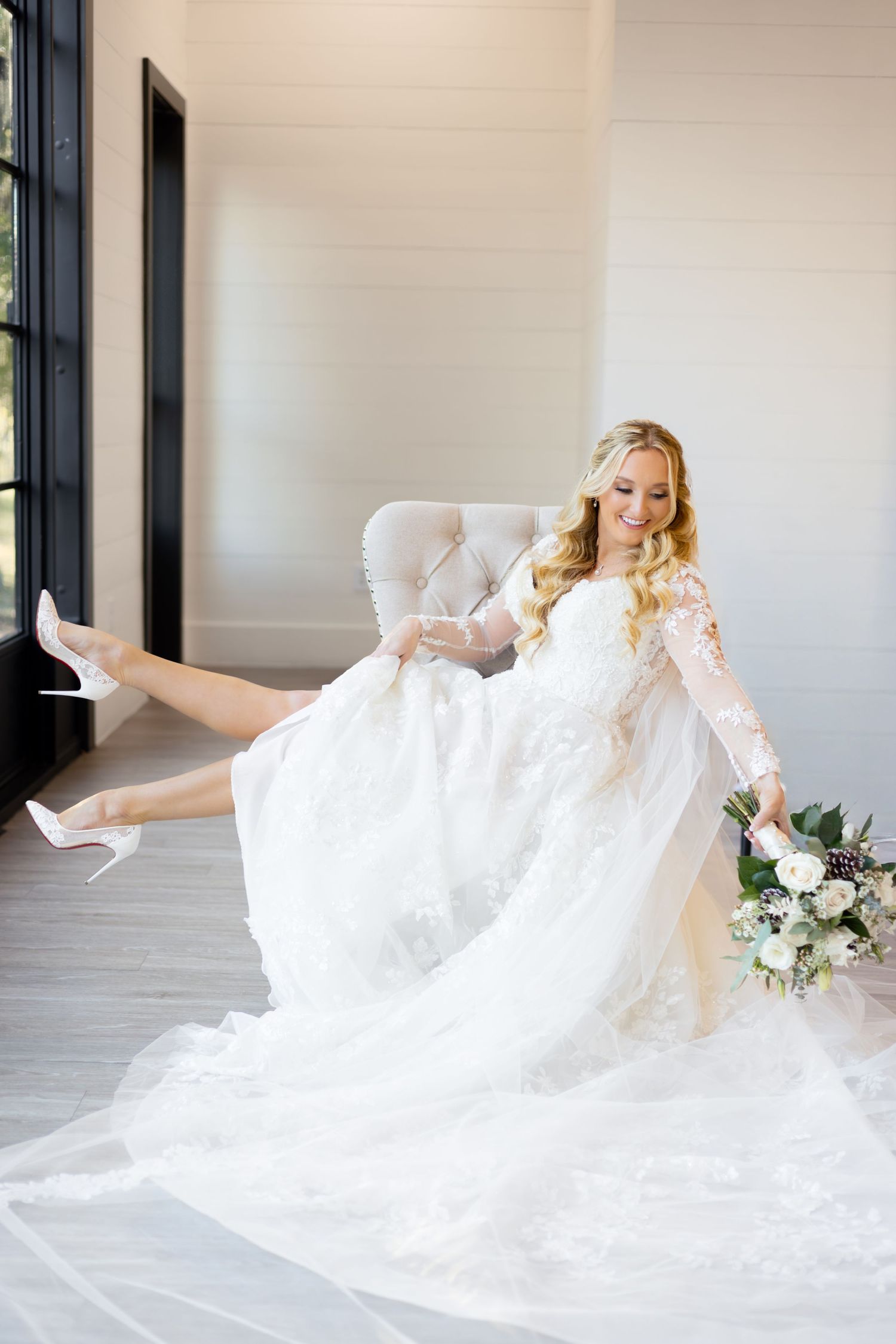 Smiling bride in lace long-sleeve wedding gown sits in chair, holding bouquet and showing white heels.