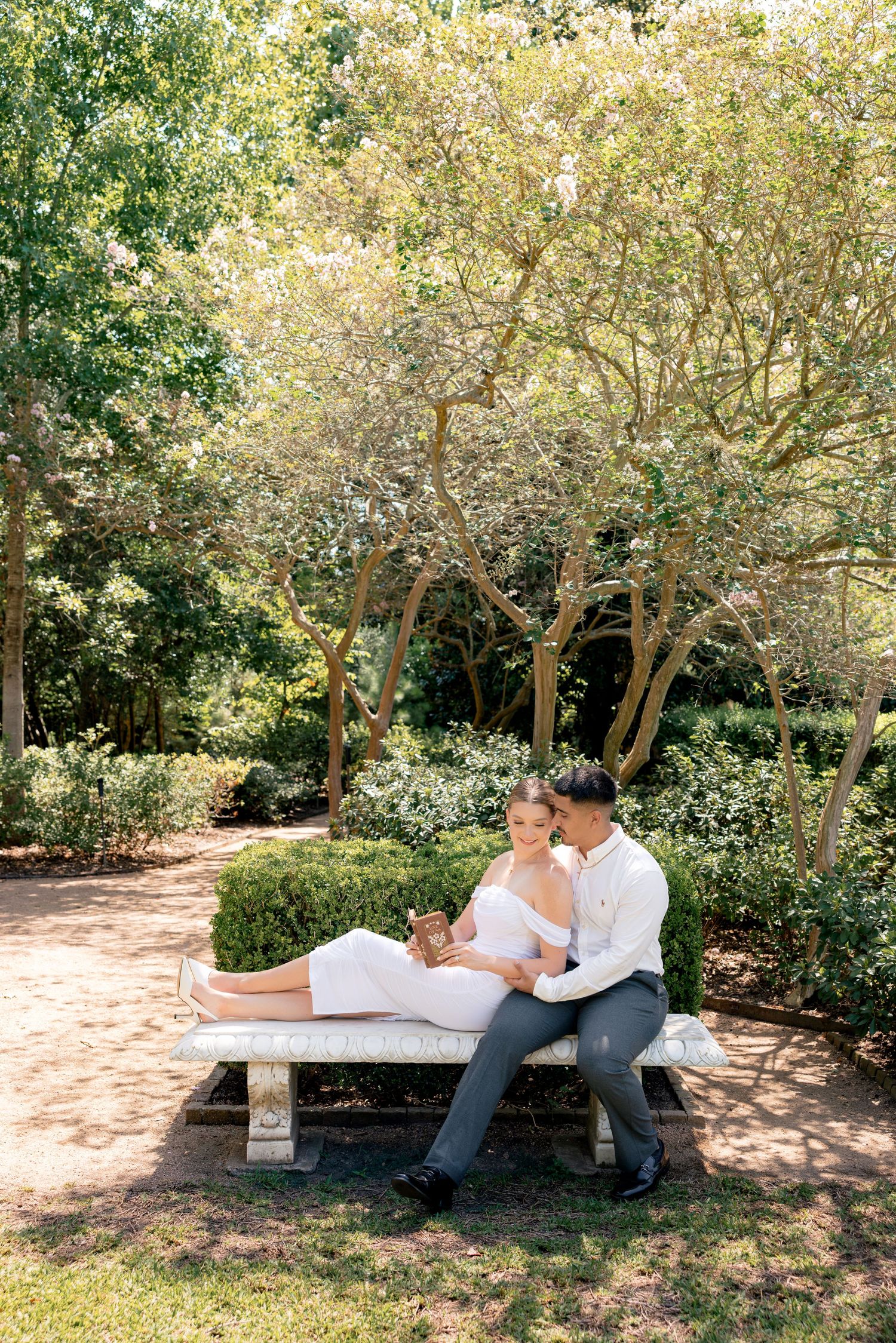 Couple sitting on a stone bench in a lush garden, woman in white dress resting legs on mans lap.