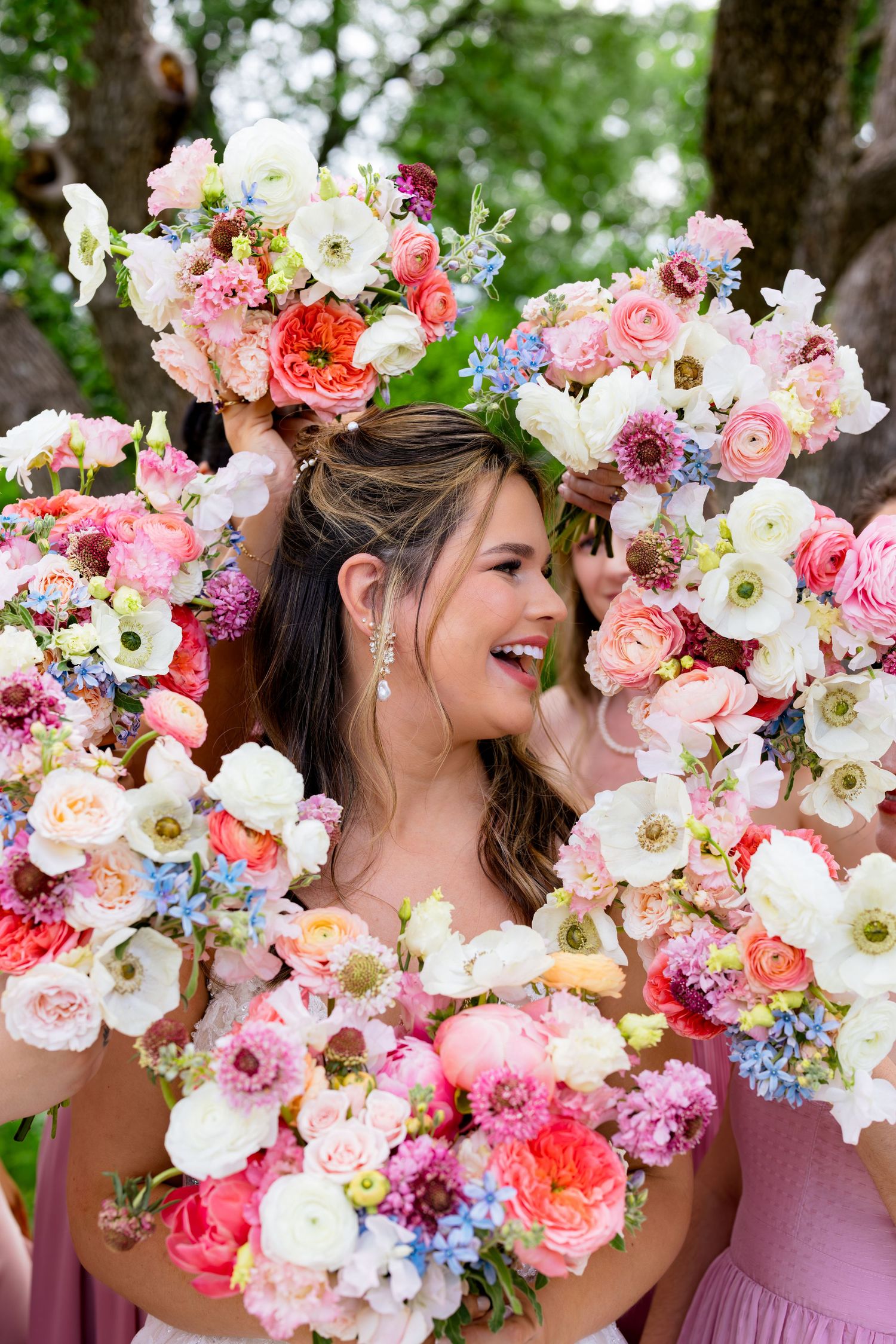 Bride laughing surrounded by colorful pink, white, and coral wedding bouquets held by bridesmaids outdoors.