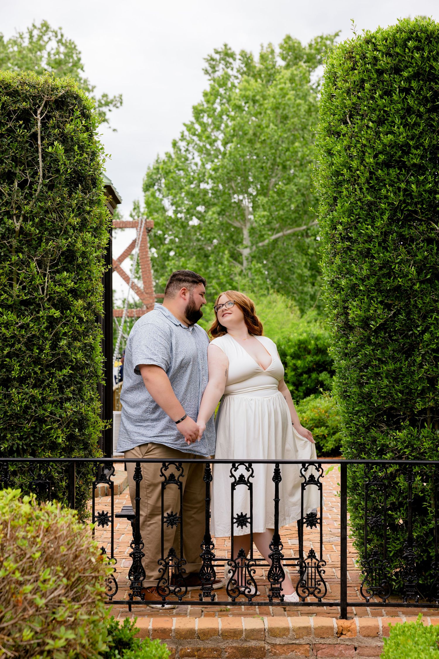 Couple holding hands and gazing at each other in a lush garden with tall hedges and ornate iron fence.