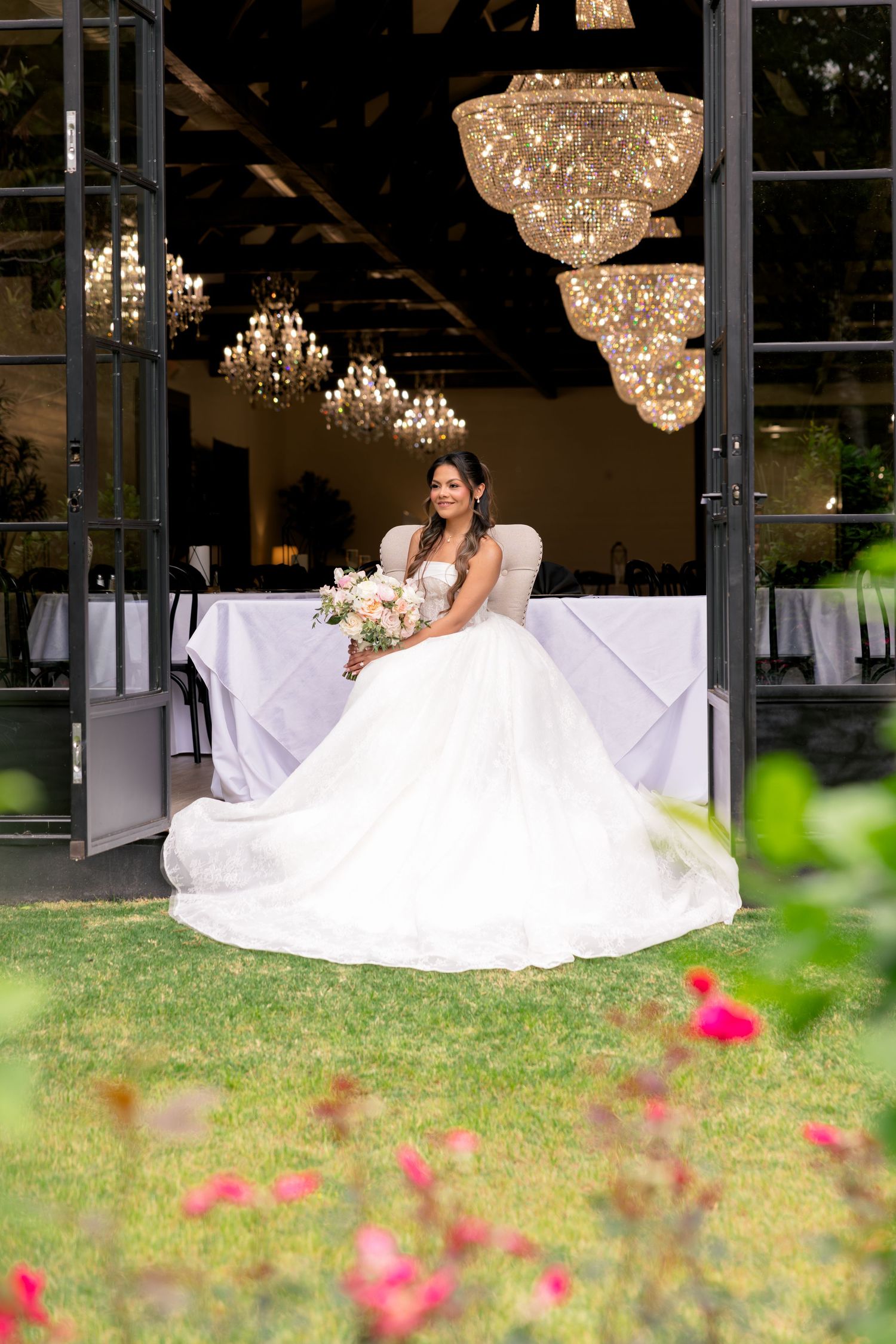 Bride in a white ballgown holding a bouquet, seated in a doorway with chandeliers and reception tables behind her.