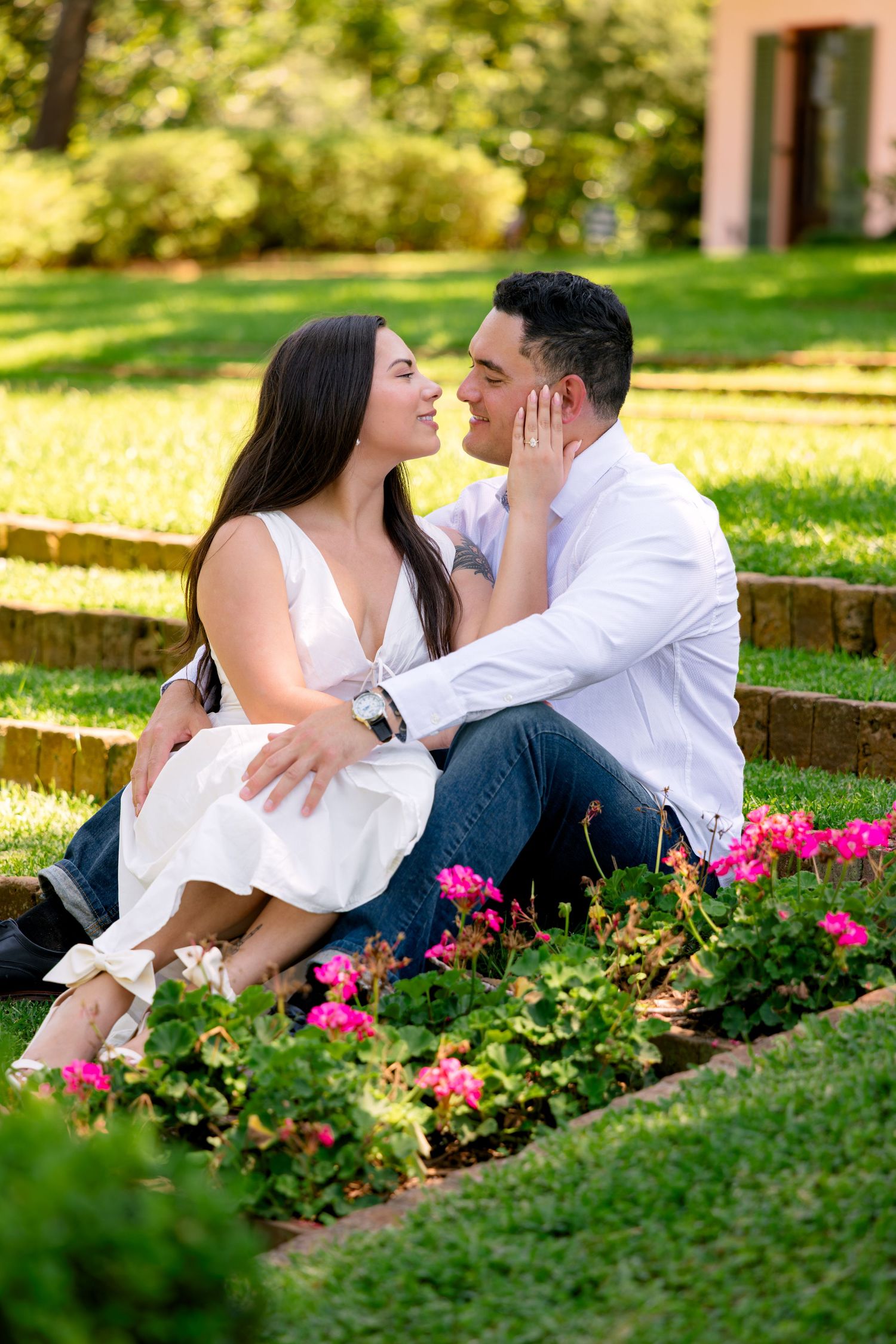 A couple shares a romantic kiss while sitting among pink flowers in a lush green garden park setting.