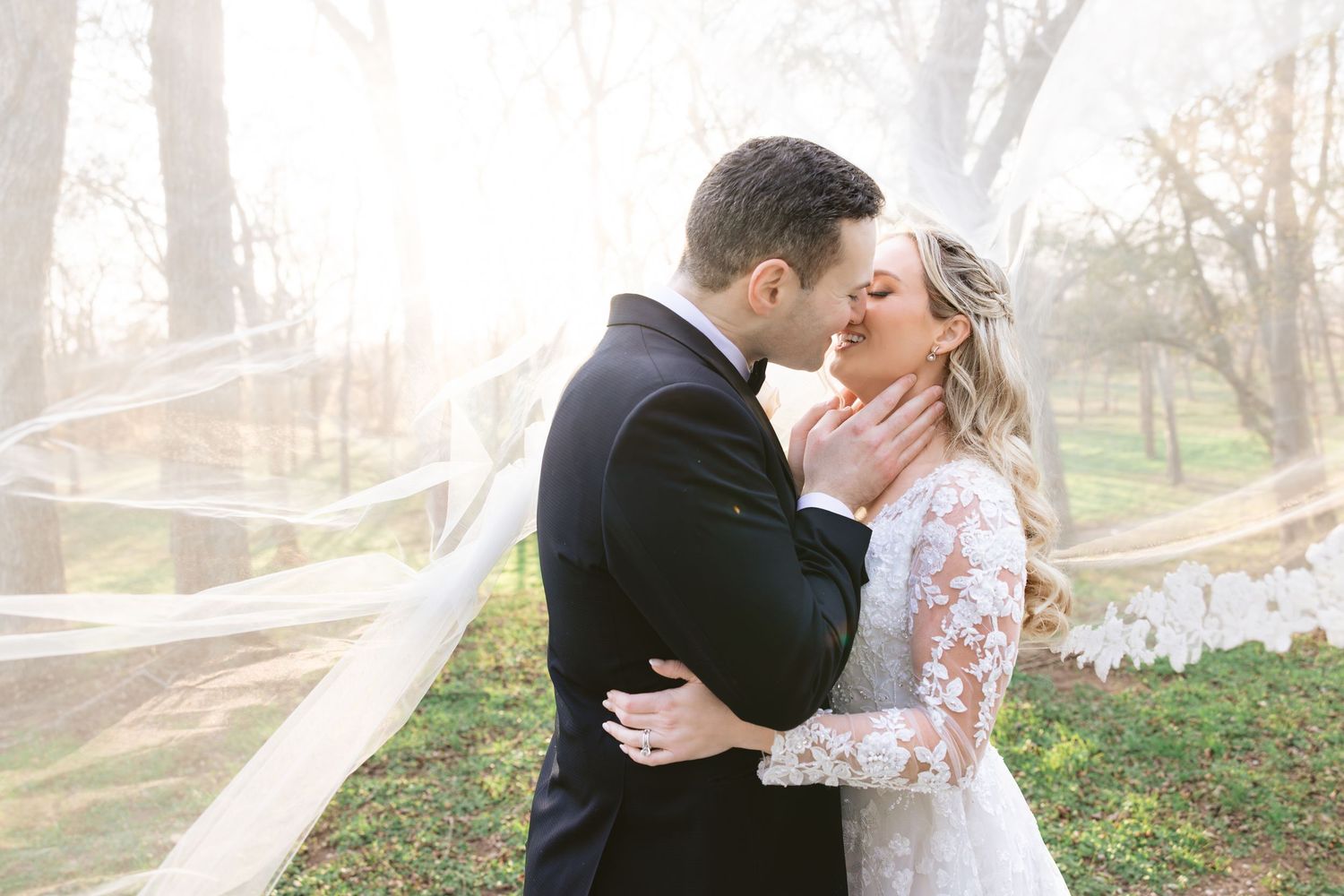 Bride and groom share a tender almost-kiss outdoors, with a flowing veil caught in the breeze in warm sunlight.