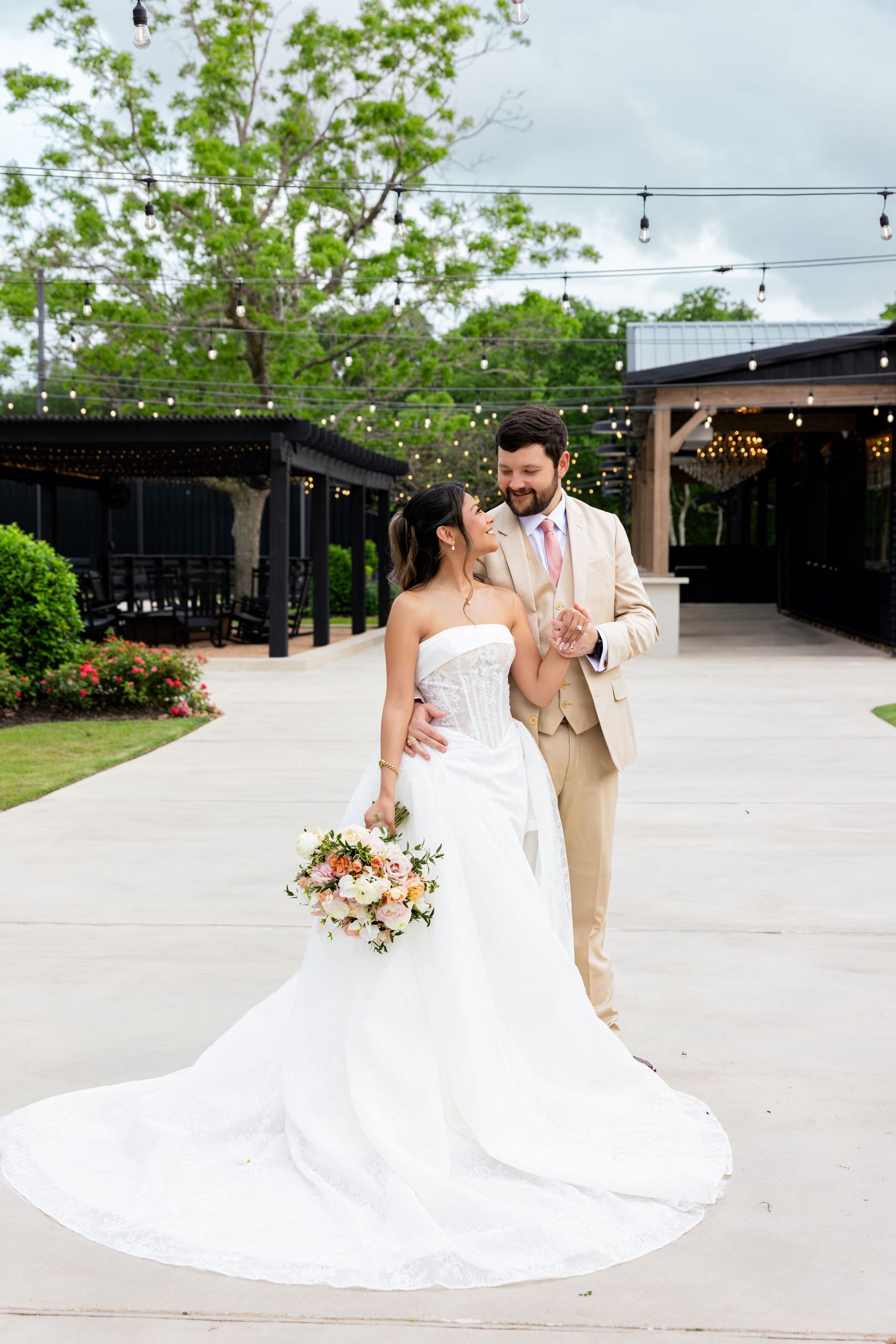 Bride in strapless white gown holding bouquet walks with groom in tan suit on outdoor wedding venue patio.
