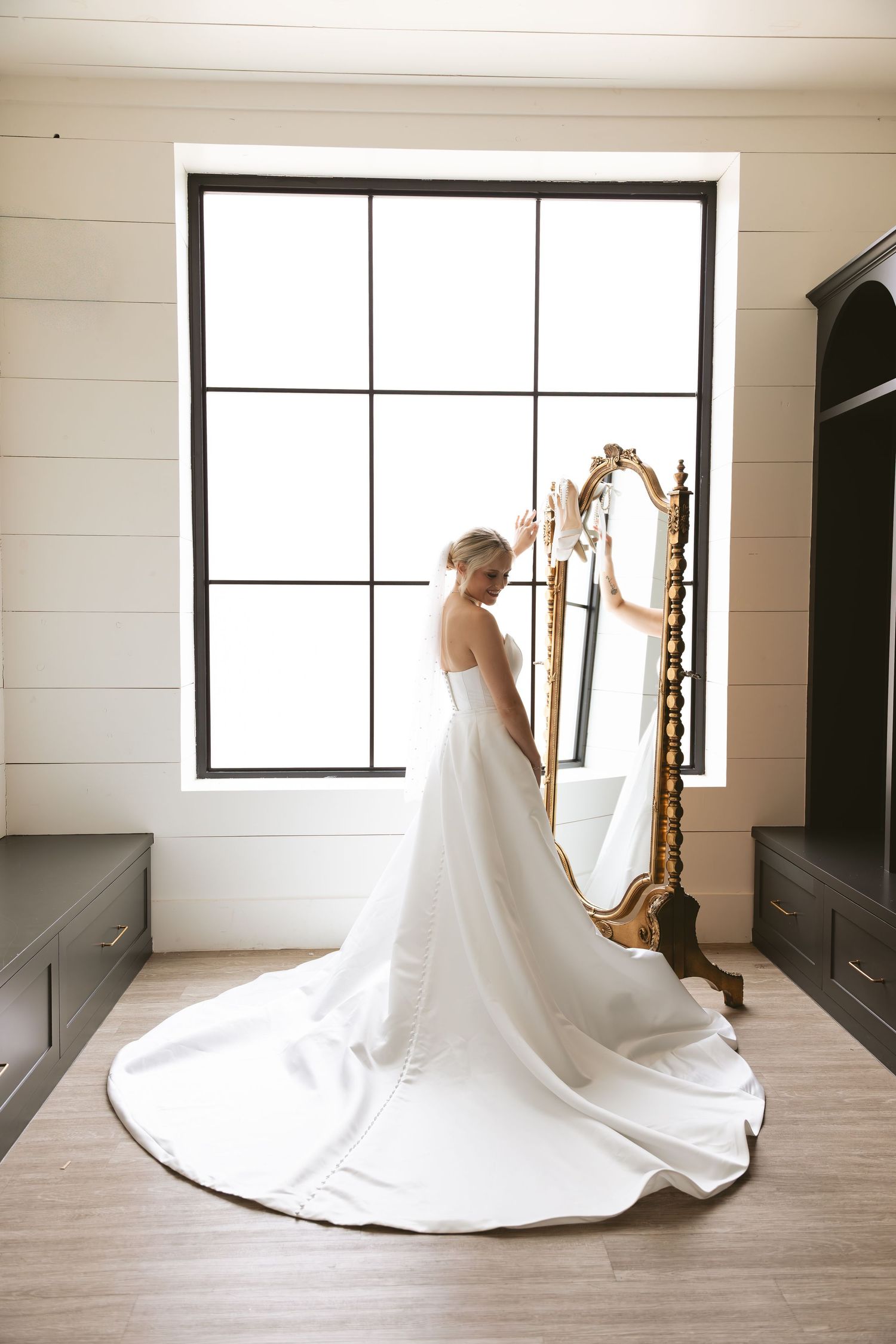 Bride in elegant white gown with long train stands before ornate gold mirror near large window.