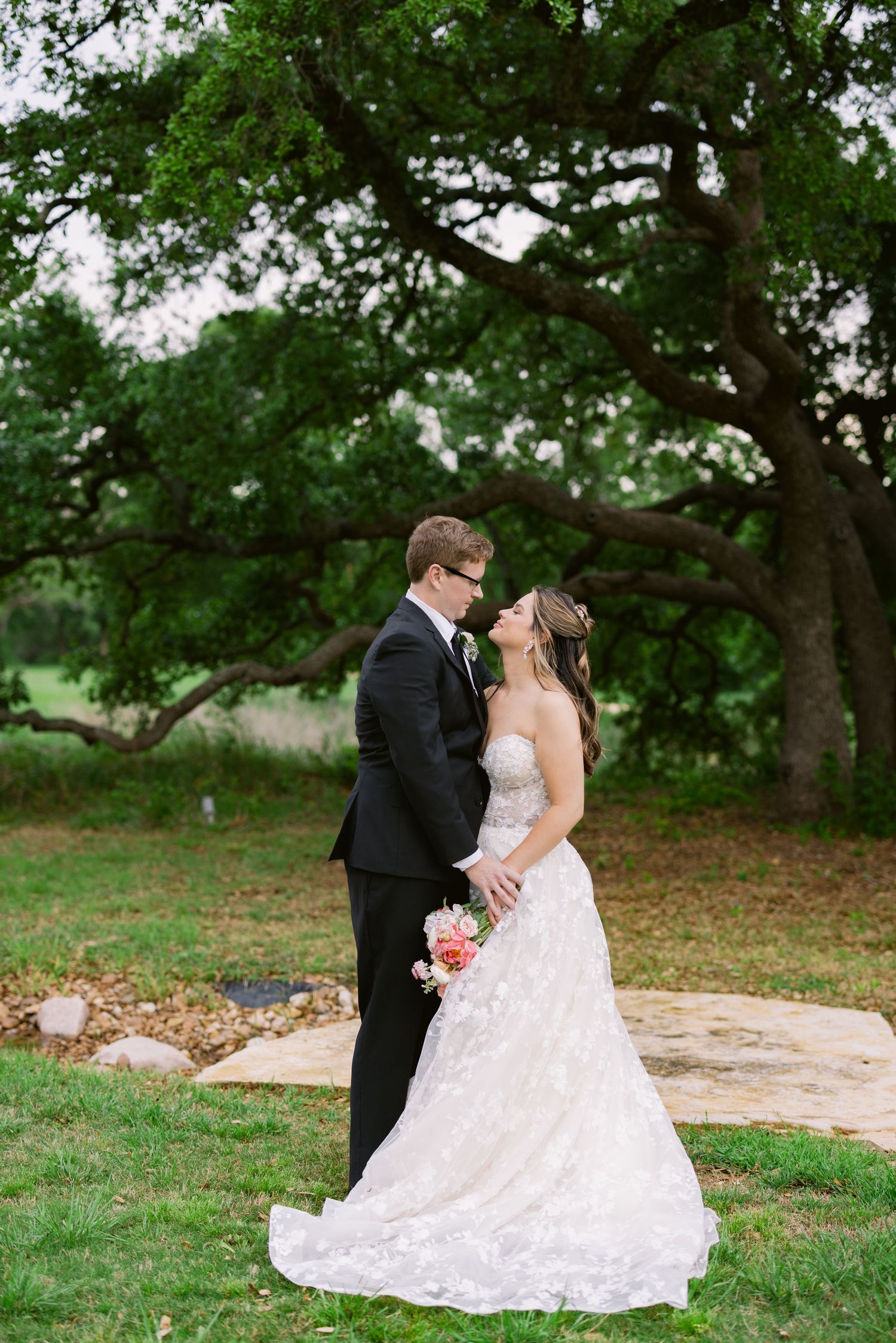 Bride and groom share a romantic moment under a large oak tree, bride in lace gown holding a floral bouquet.
