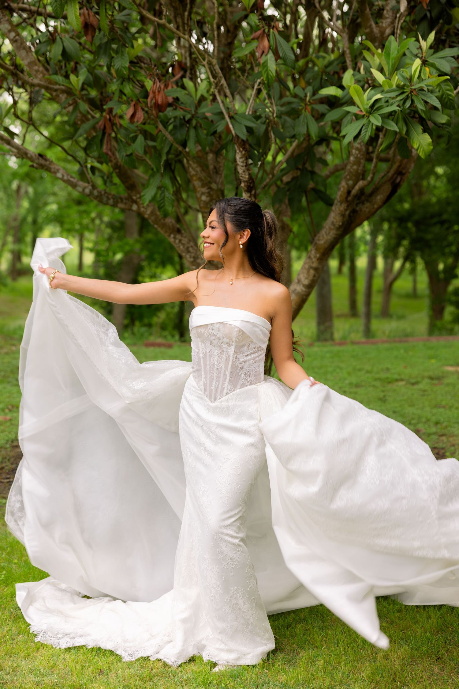 Bride in white strapless mermaid gown twirling veil outdoors in lush green garden setting.