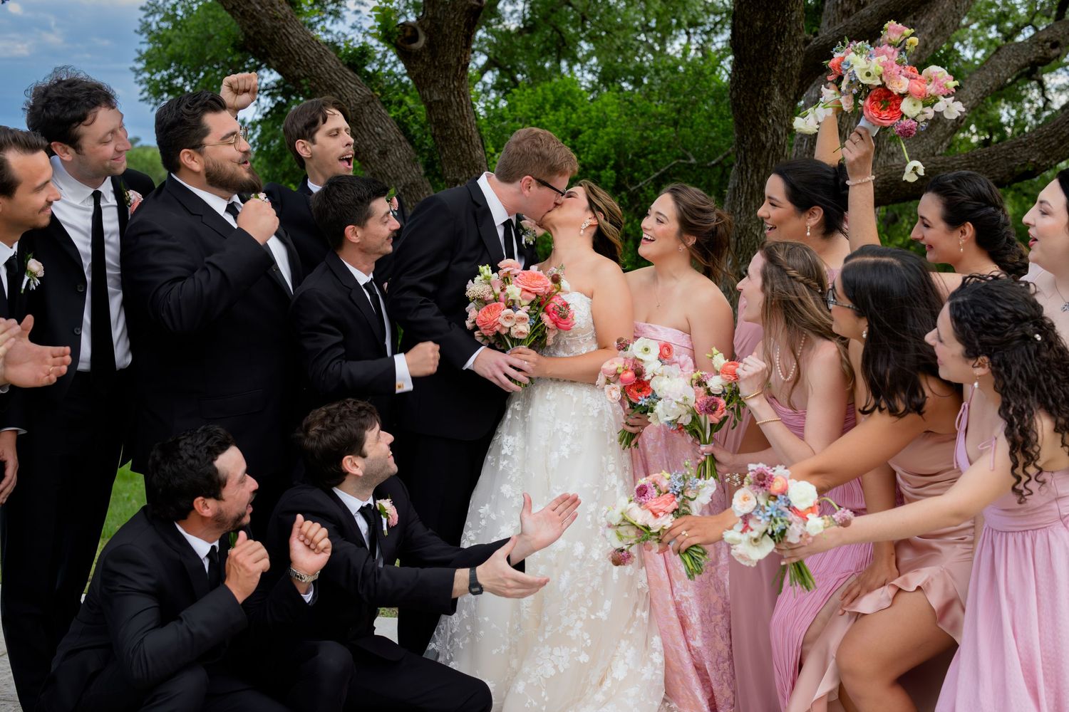 Wedding party celebrates newlyweds kissing, groomsmen in black suits and bridesmaids in pink dresses holding colorful bouquets outdoors.