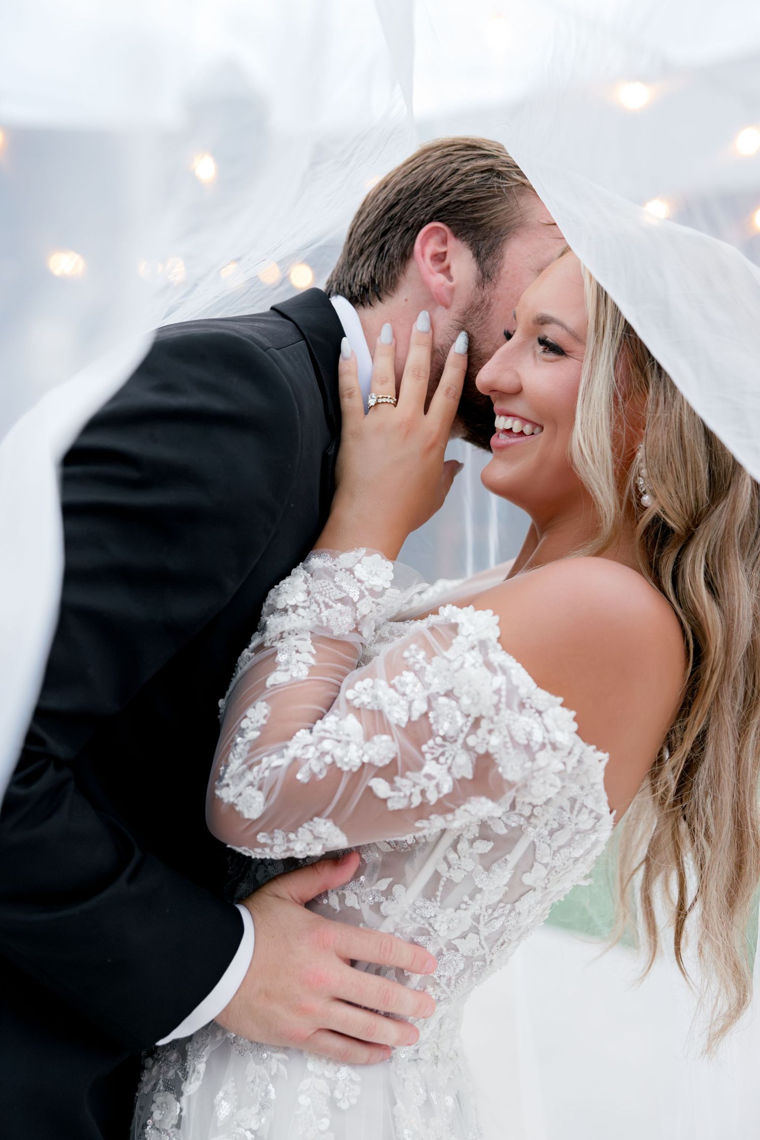 Bride and groom share romantic moment under veil, bride smiling in lace off-shoulder gown with bokeh lights behind them.